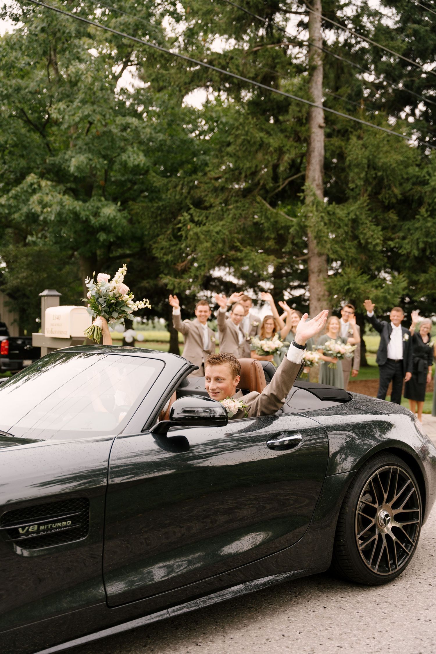 Groom waves from a black Mercedes-AMG GT convertible as wedding party celebrates in the background.