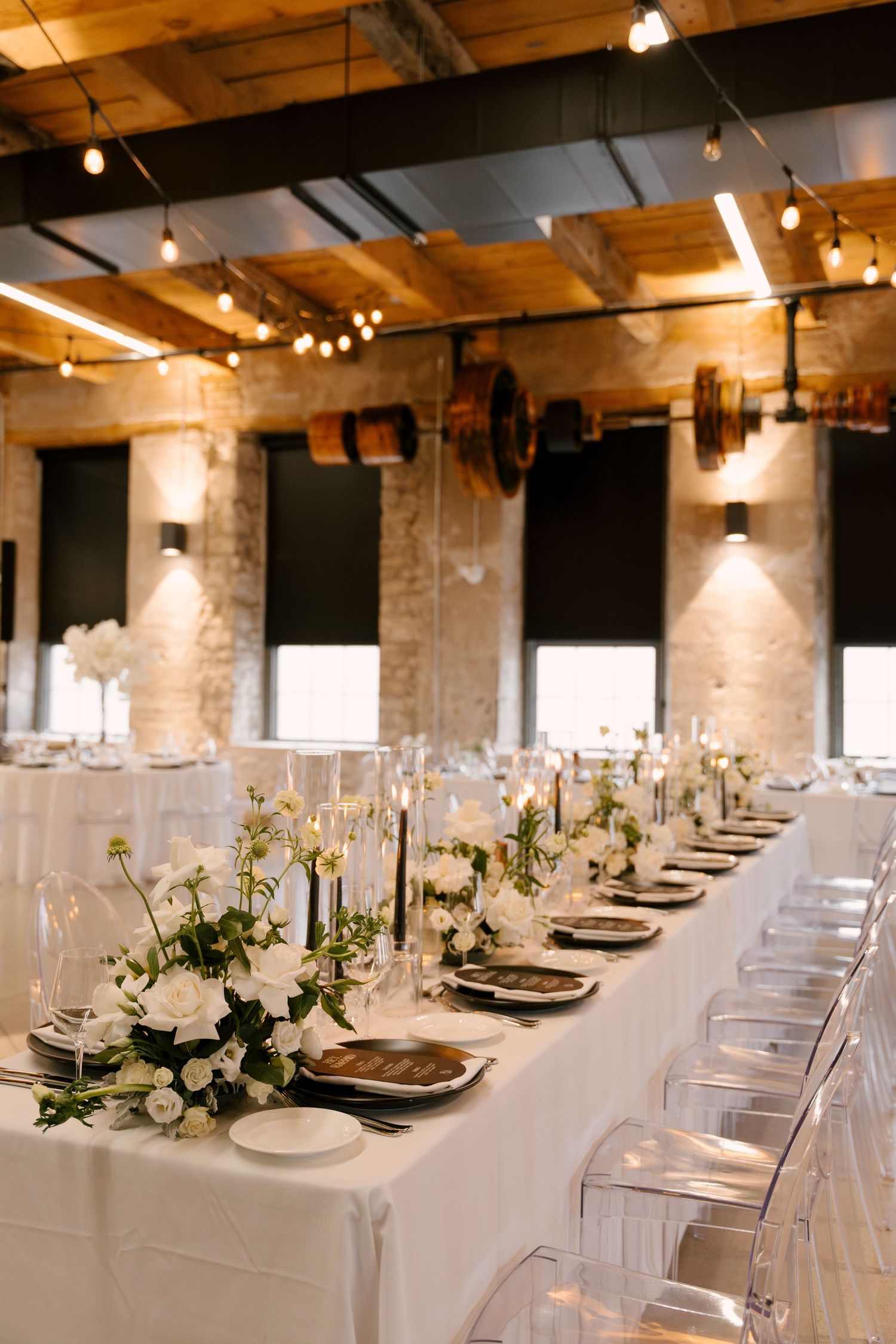Elegant wedding reception with long white tables, black candles, white floral centerpieces, and ghost chairs in an industrial venue.