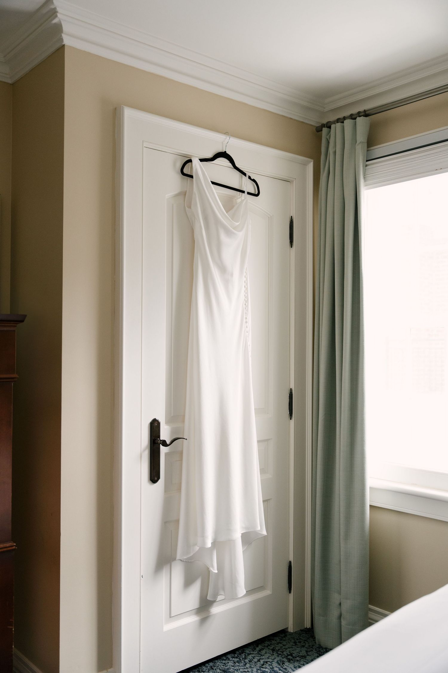 Elegant white bridal gown hanging on door hanger in softly lit hotel room before wedding.