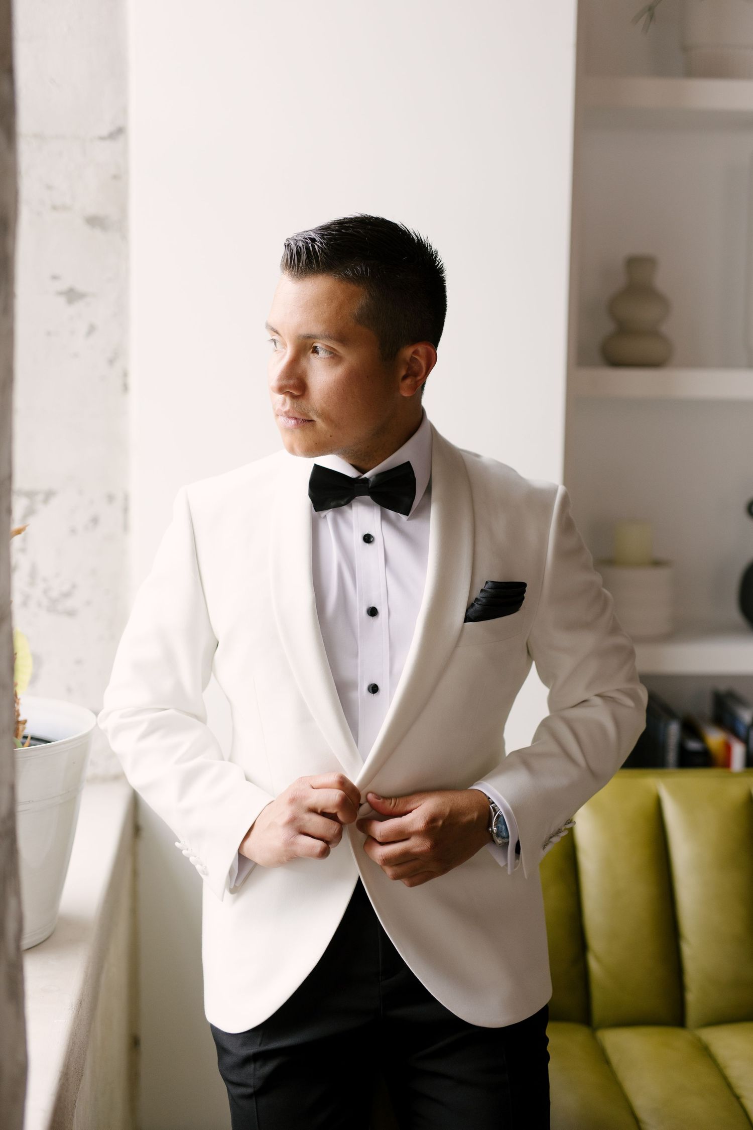 A dapper groom in a white tuxedo jacket and black bow tie buttoning his jacket while gazing out a window.