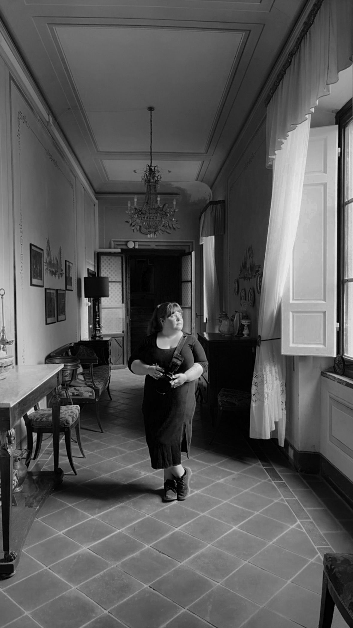 Black and white photo of woman in black dress walking through ornate vintage corridor with chandelier.