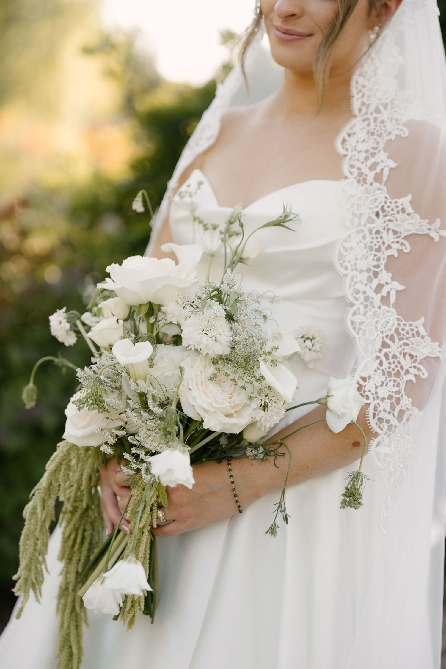 Bride in a white strapless gown holds a lush white floral bouquet with lace veil detail.