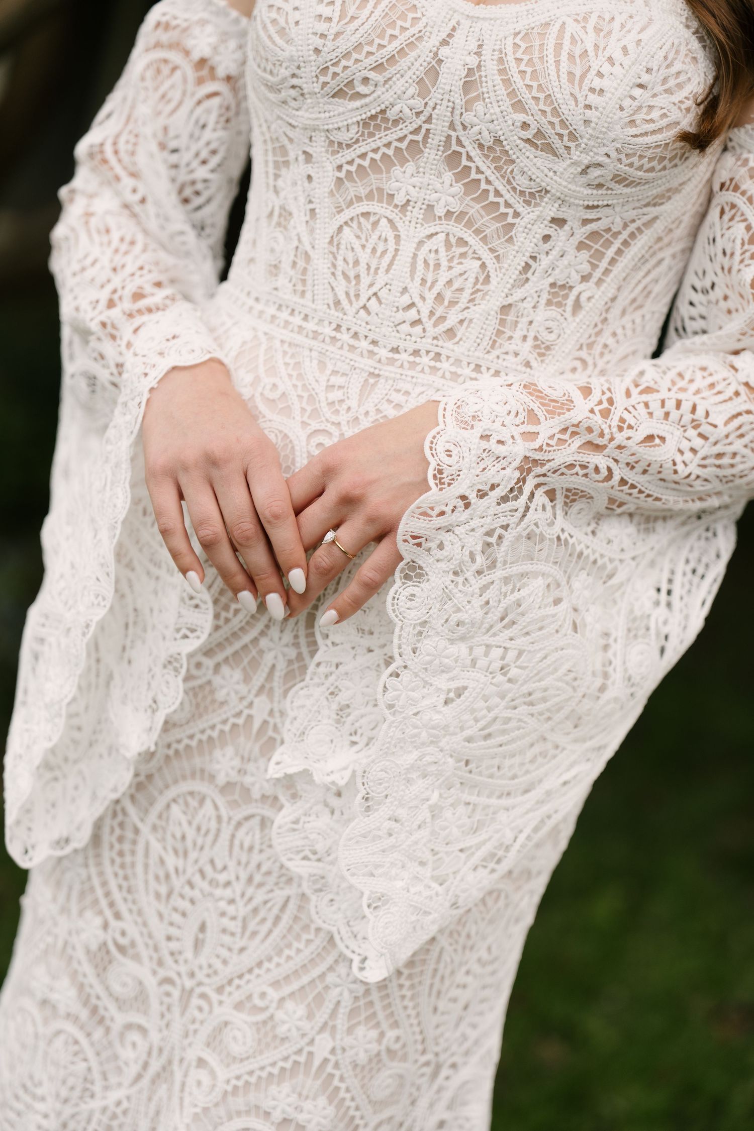 Close-up of a bride in an intricate white lace long-sleeve wedding gown, hands folded showing a diamond ring.
