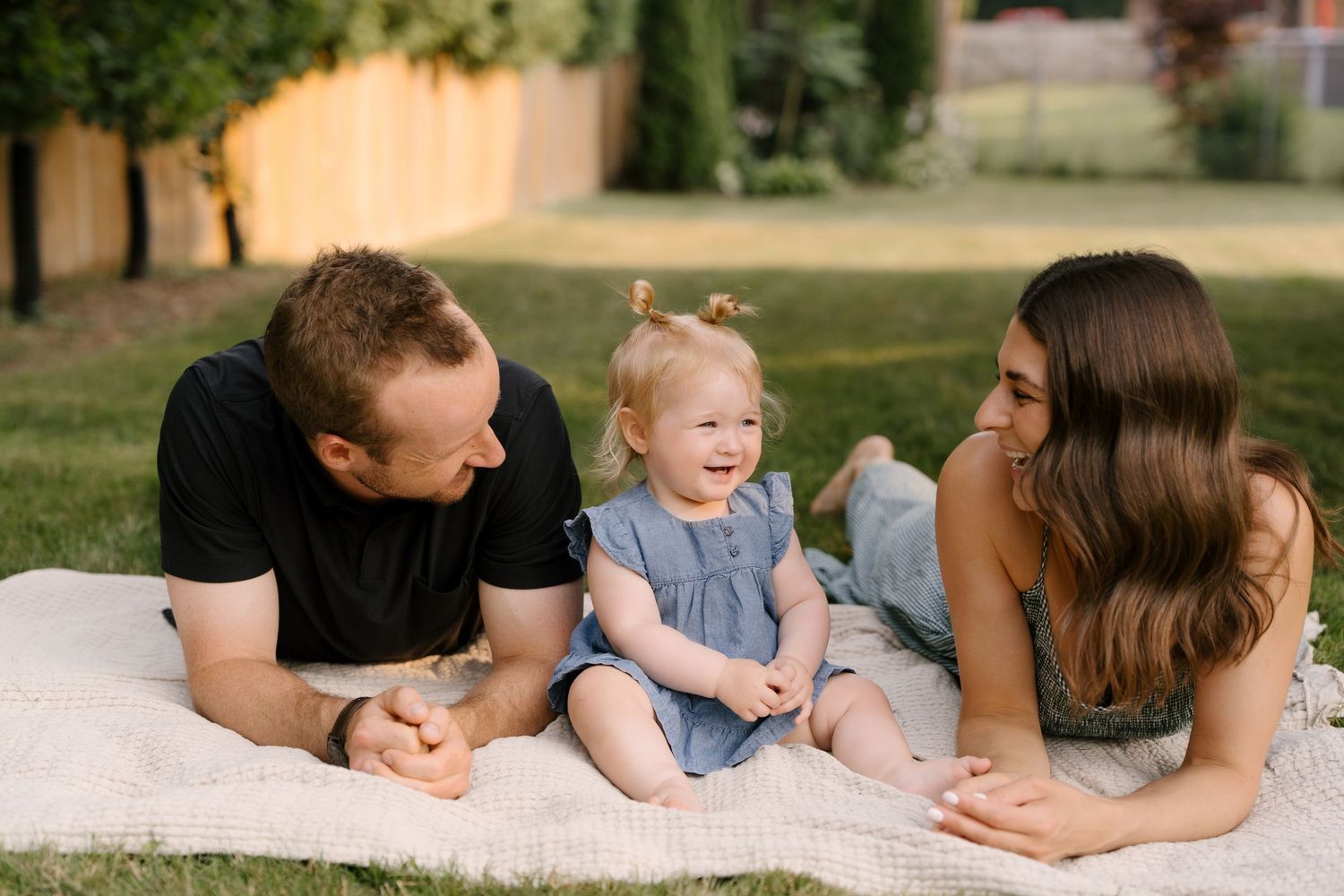 A happy family lies on a blanket in a green backyard, smiling at their cheerful toddler in blue.