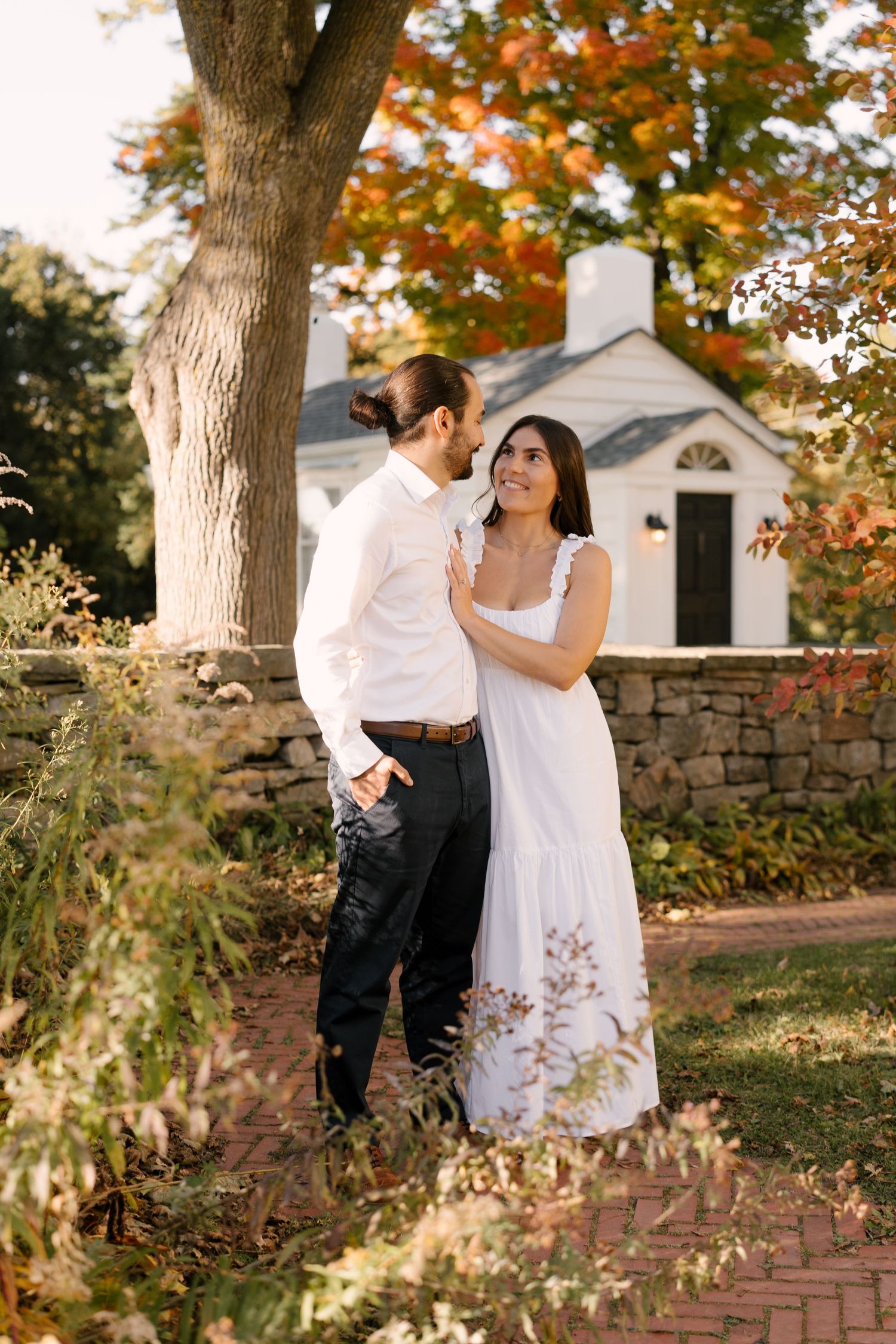 Couple sharing a tender moment in a sunlit autumn garden near a white cottage.