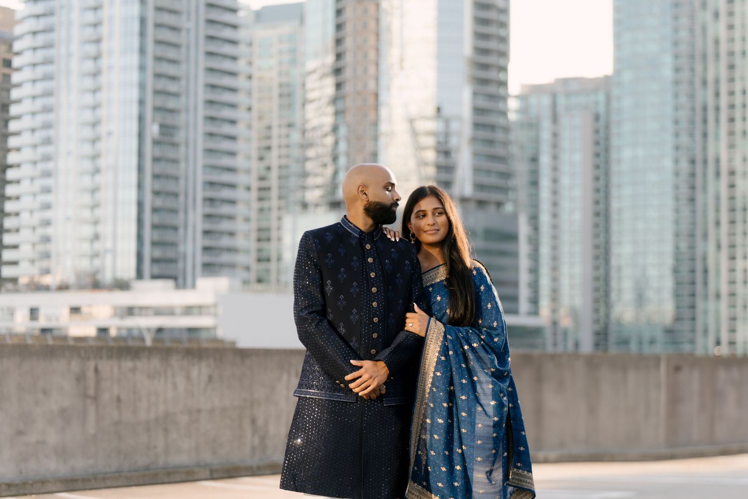 Indian couple in coordinating blue traditional outfits pose together against city skyline.