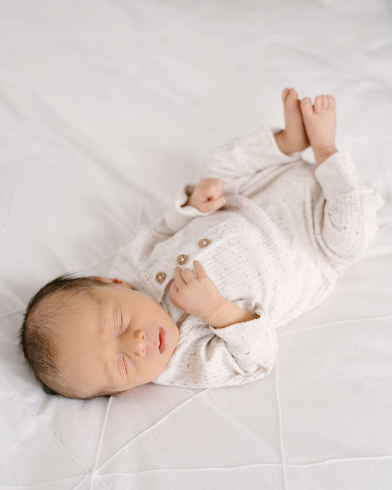 Peaceful sleeping newborn baby in cream knit romper lying on white bedding with legs raised.