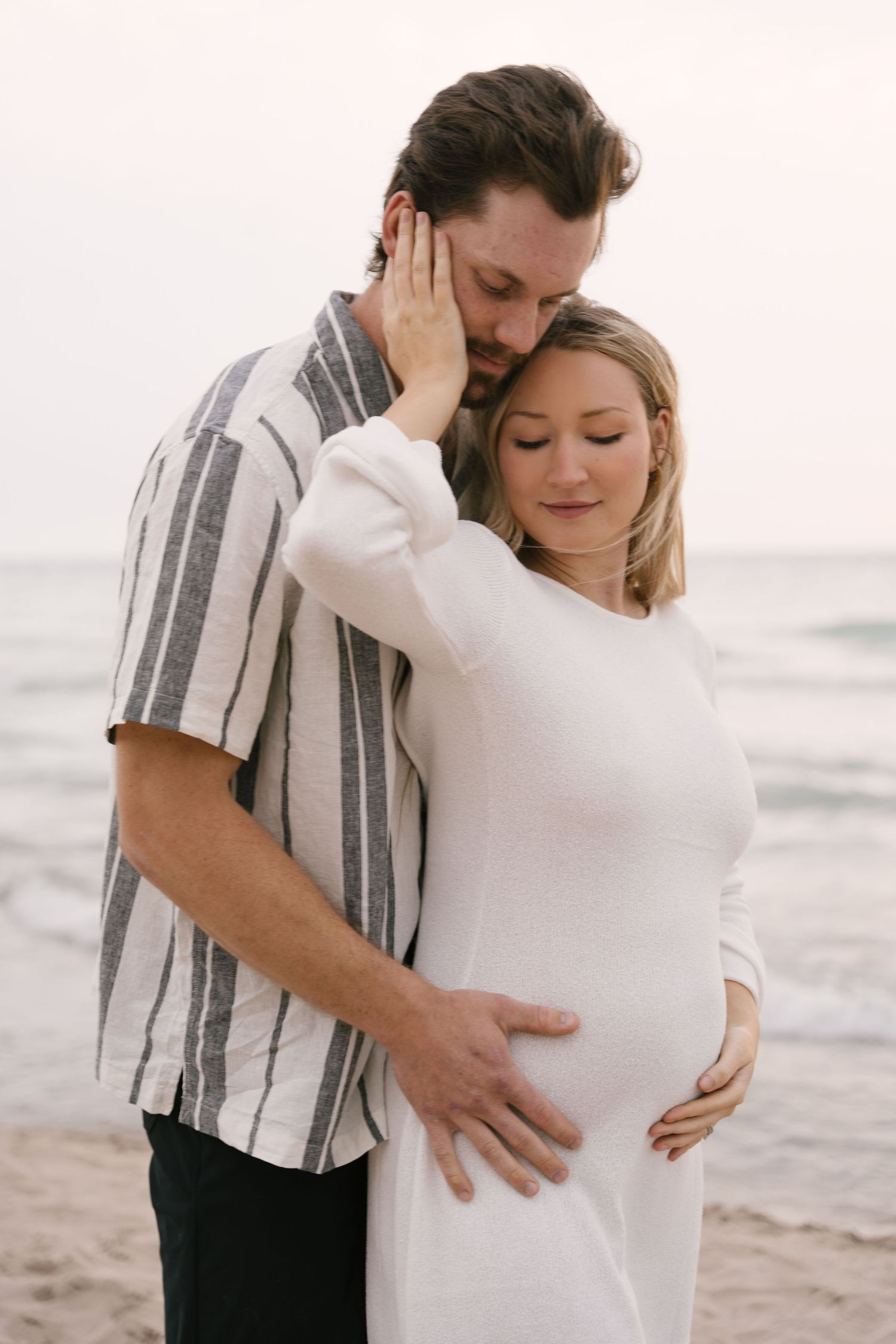 Couple embracing on beach at sunset, man kissing pregnant womans head while holding her bump.