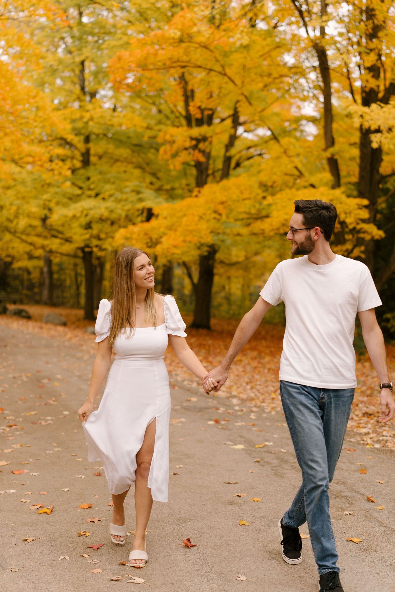 A smiling couple walks hand-in-hand along a path surrounded by brilliant fall foliage.