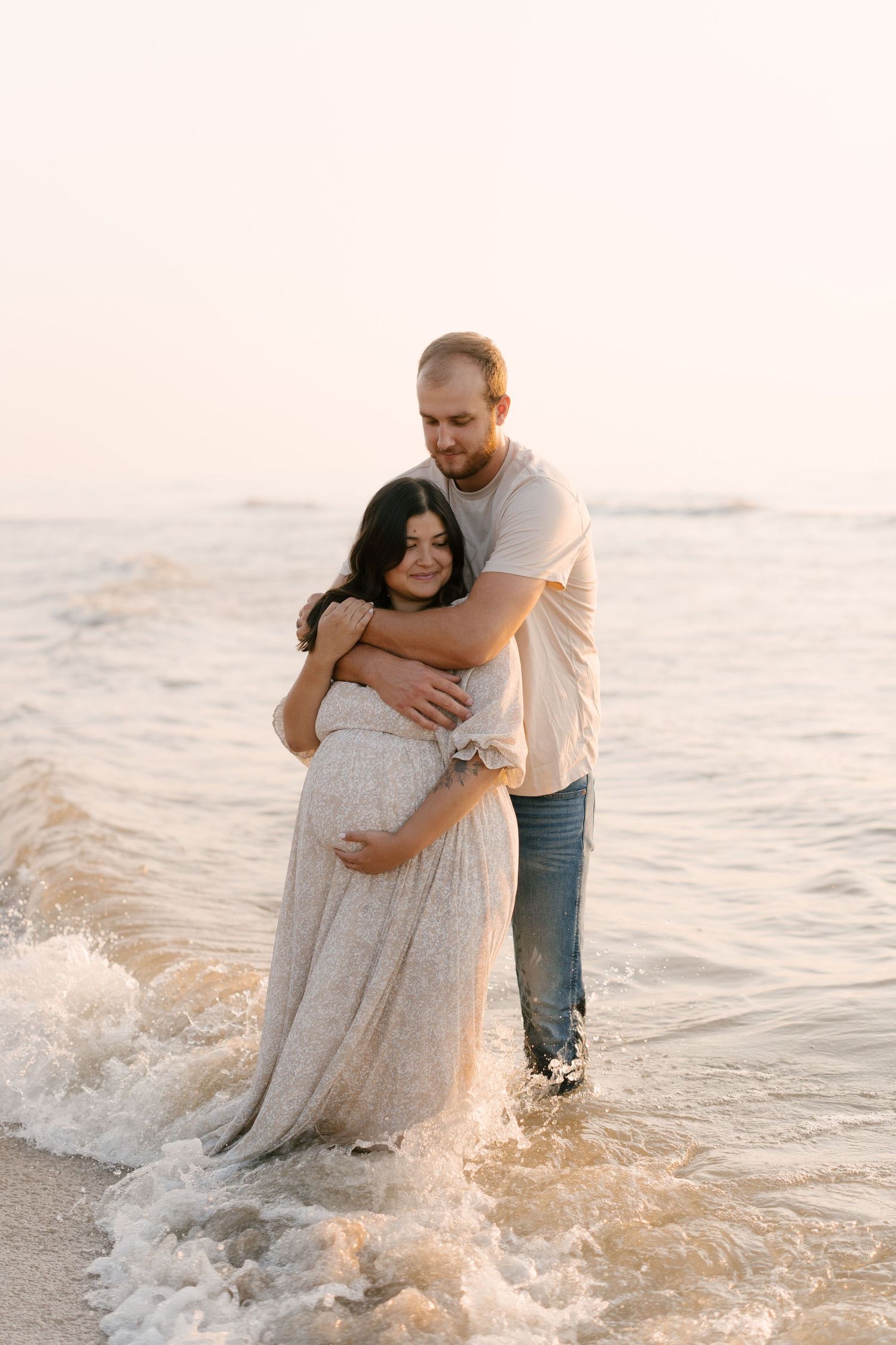 Man embracing pregnant partner from behind in ocean waves during golden hour beach photoshoot.