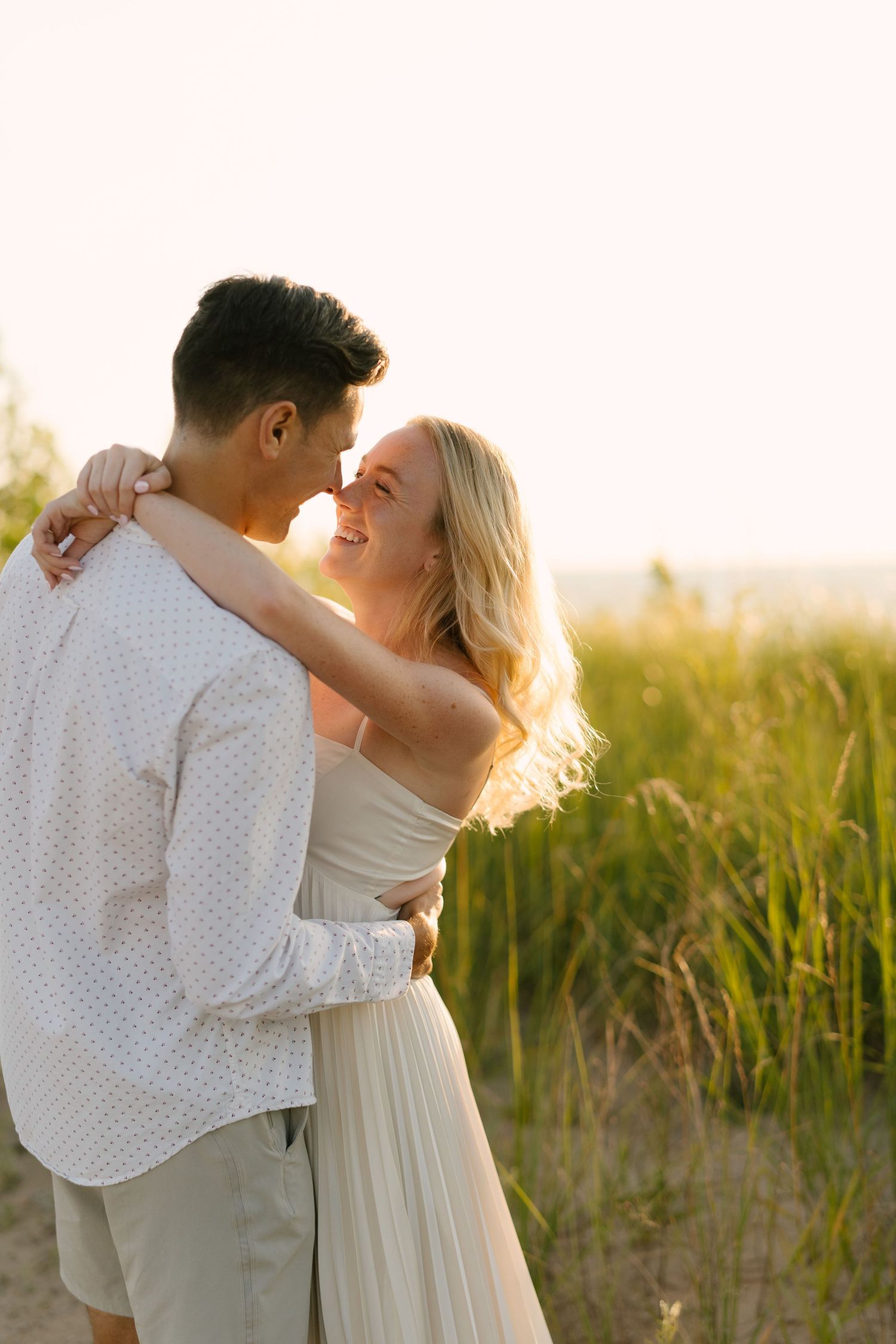Smiling blonde woman in white dress embracing man in light shirt at sunset near tall beach grass.