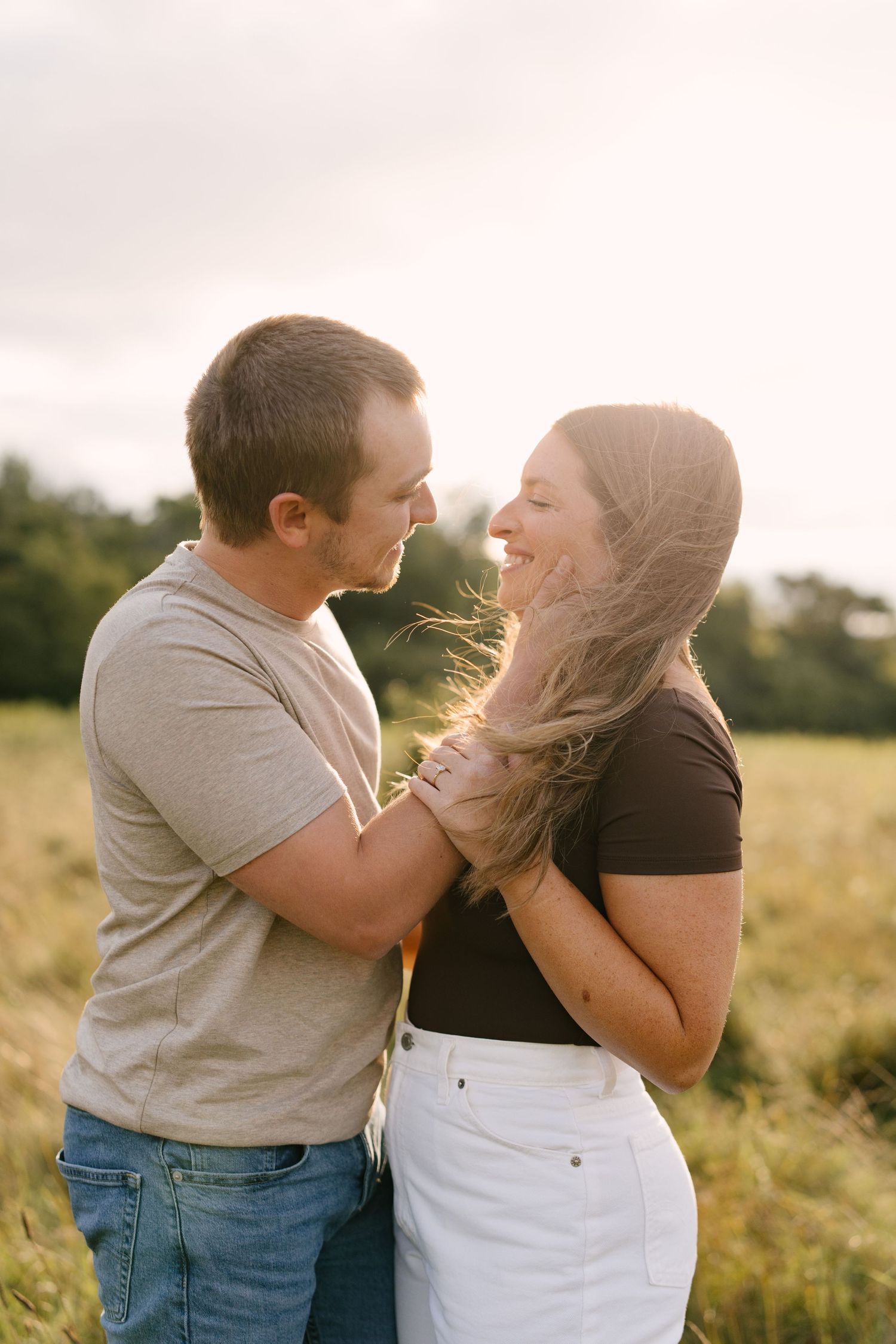 Couple facing each other laughing in a sunlit field with wind blowing through her hair.