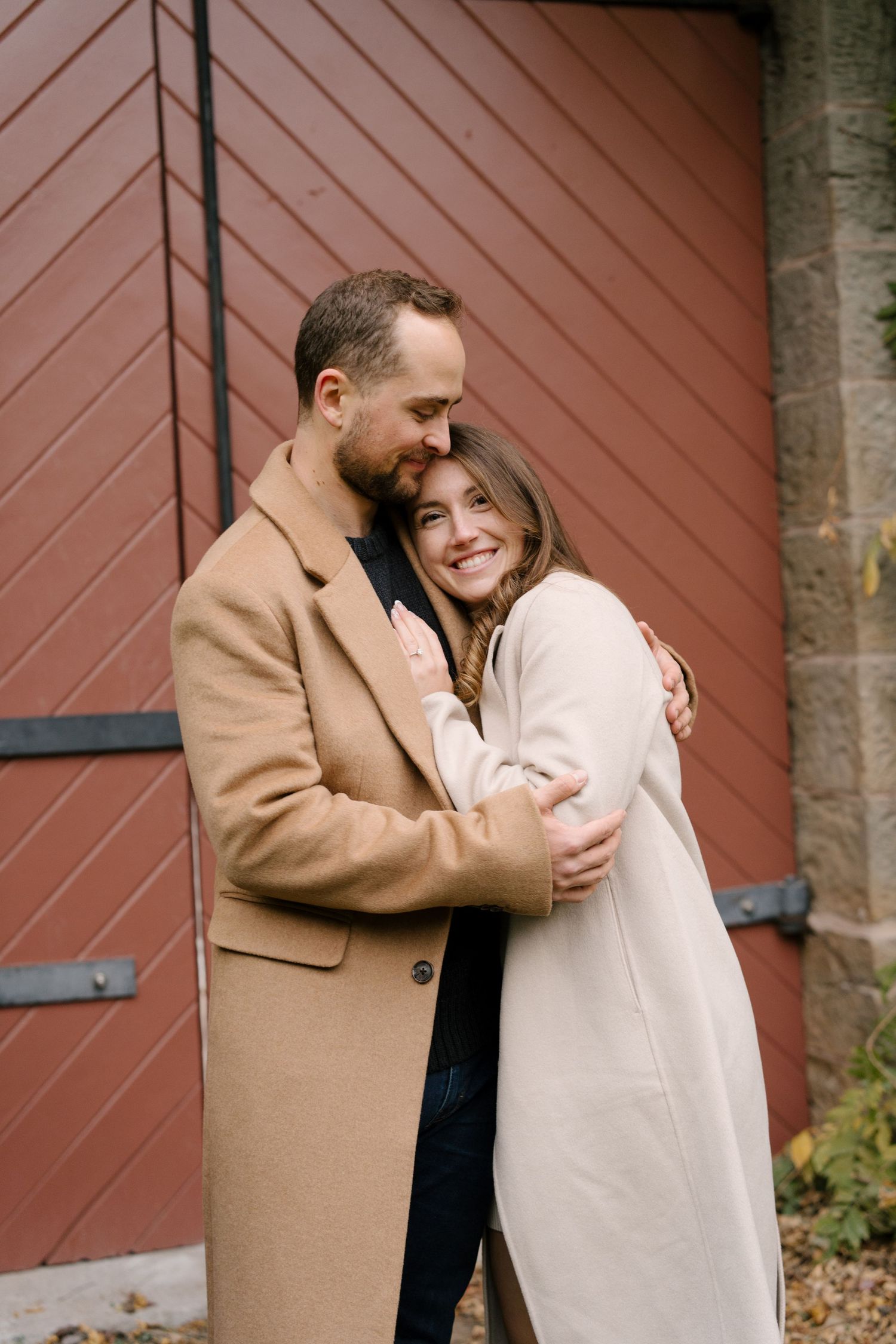 Happy couple embracing and smiling before a red barn wall in matching beige and cream autumn coats.