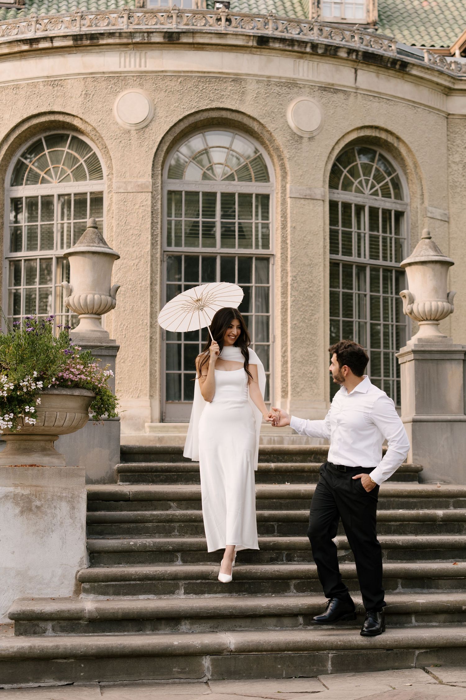 Bride descending grand stone steps holding groom's hand outside elegant historic building with arched windows.