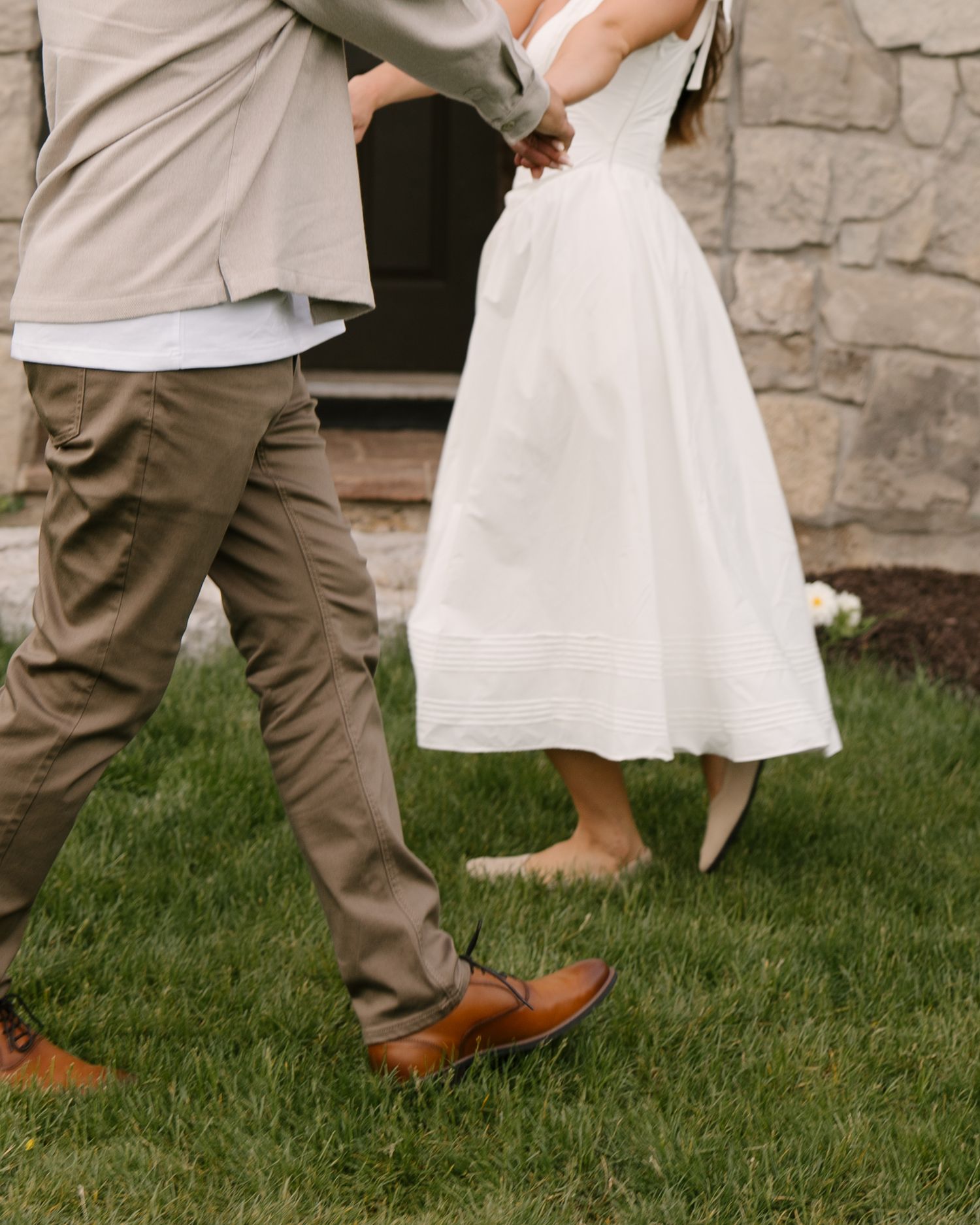 Couple walking hand-in-hand on green grass, woman in white midi dress, man in tan jacket and brown shoes.