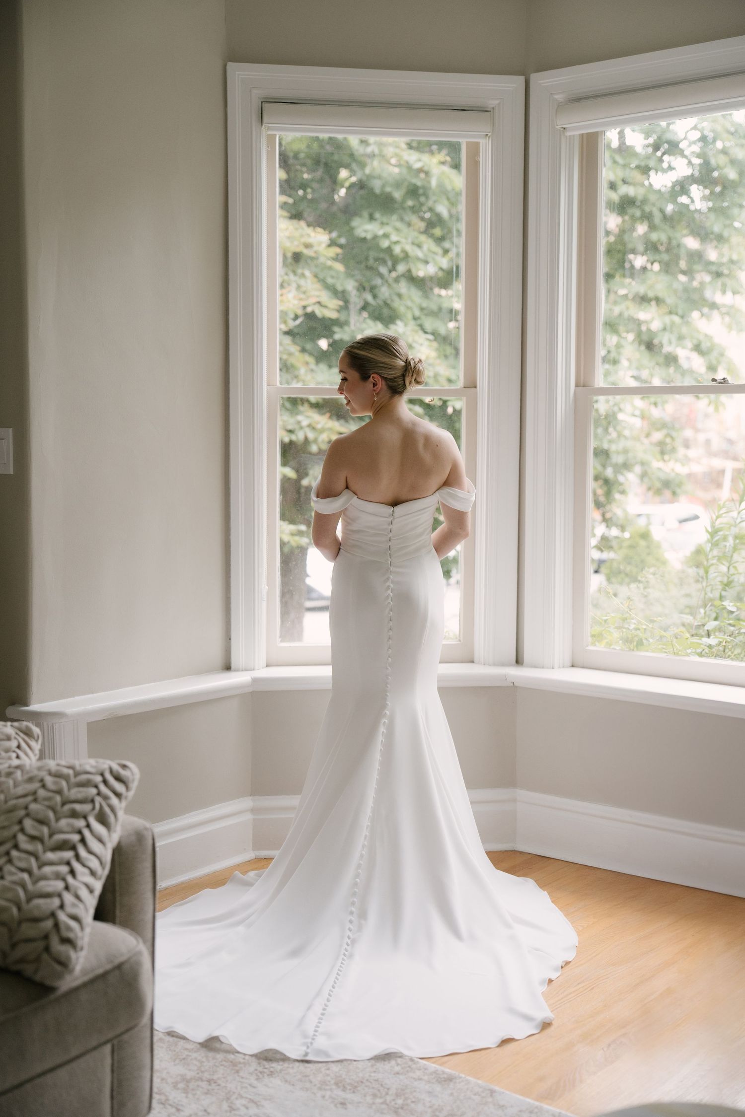 Bride in elegant off-shoulder white mermaid gown with train standing at bay window.