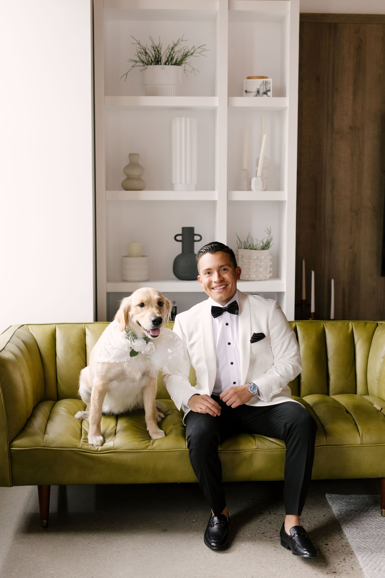 Smiling groom in white tuxedo sits beside golden retriever dog on green velvet sofa at Tapestry Hall wedding venue.