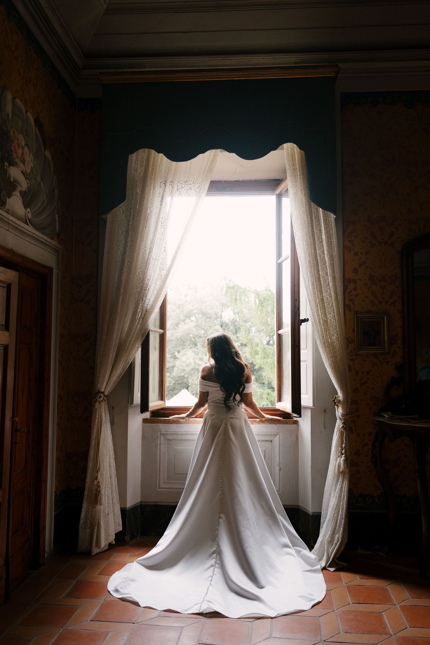 Bride in elegant white gown with train stands at ornate open window in historic villa room.