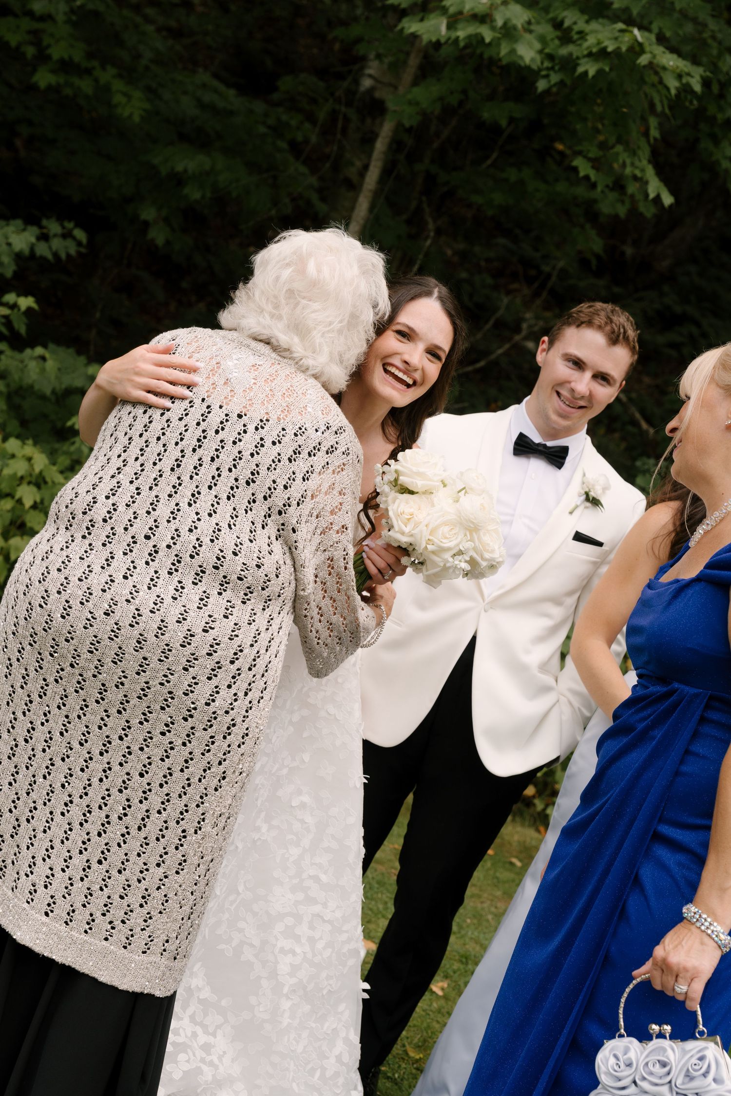 Smiling bride hugs an elderly guest while groom looks on happily during outdoor wedding reception.