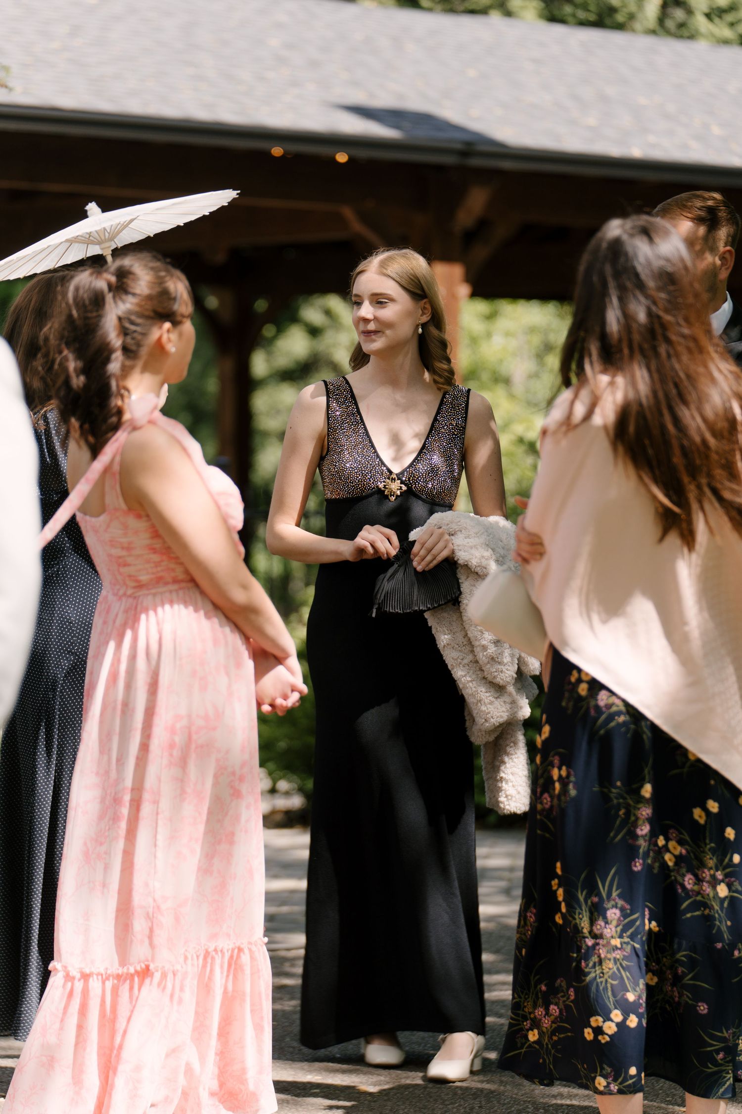 Elegantly dressed wedding guest in black gown chats with others outside rustic outdoor venue.