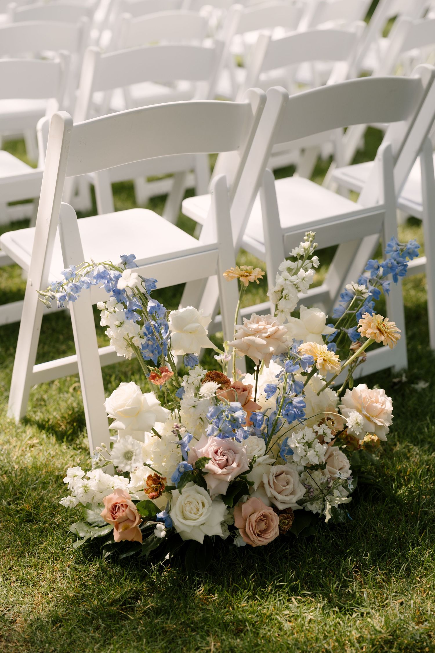 Outdoor wedding ceremony setup with white folding chairs on grass and an elegant floral arrangement of roses and blue flowers.