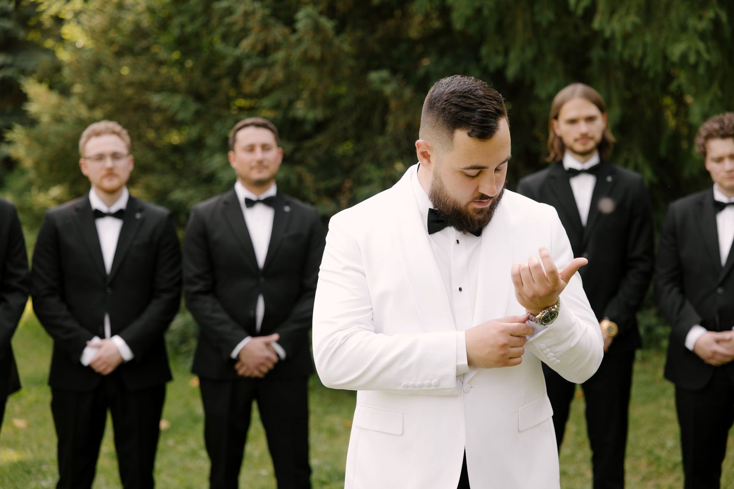 Groom in white tuxedo adjusting cufflinks while groomsmen in black tuxedos stand behind him near evergreen trees.