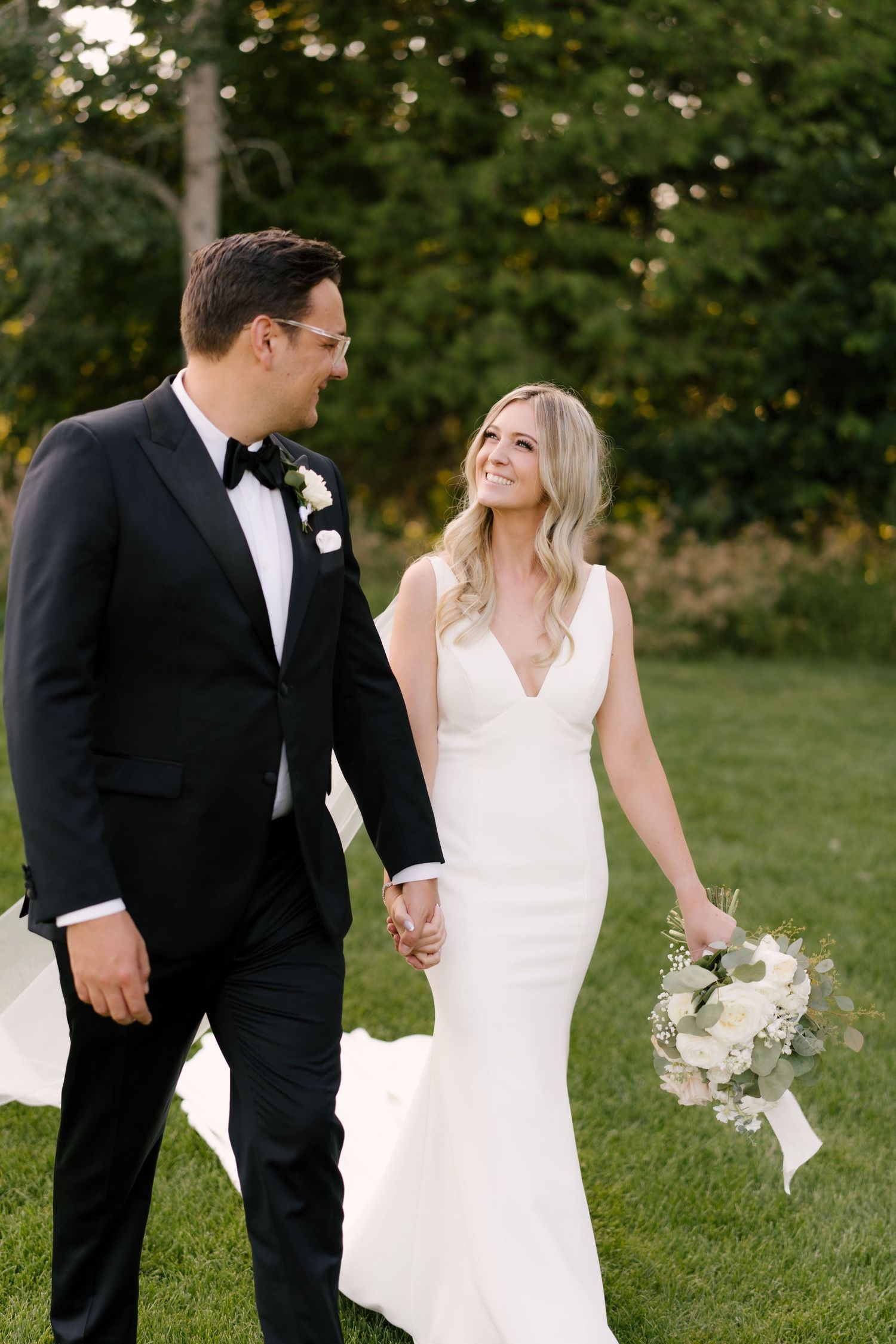 Bride in white mermaid gown and groom in black tuxedo holding hands while walking on grass.