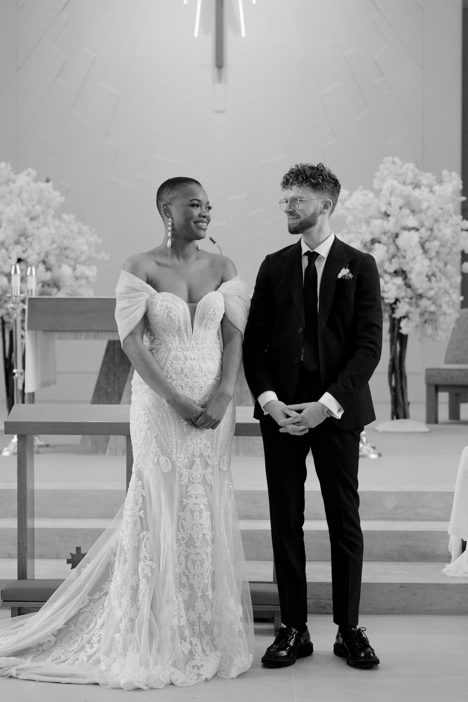 Smiling bride in lace gown and groom in black suit stand together at church altar in black and white photo.