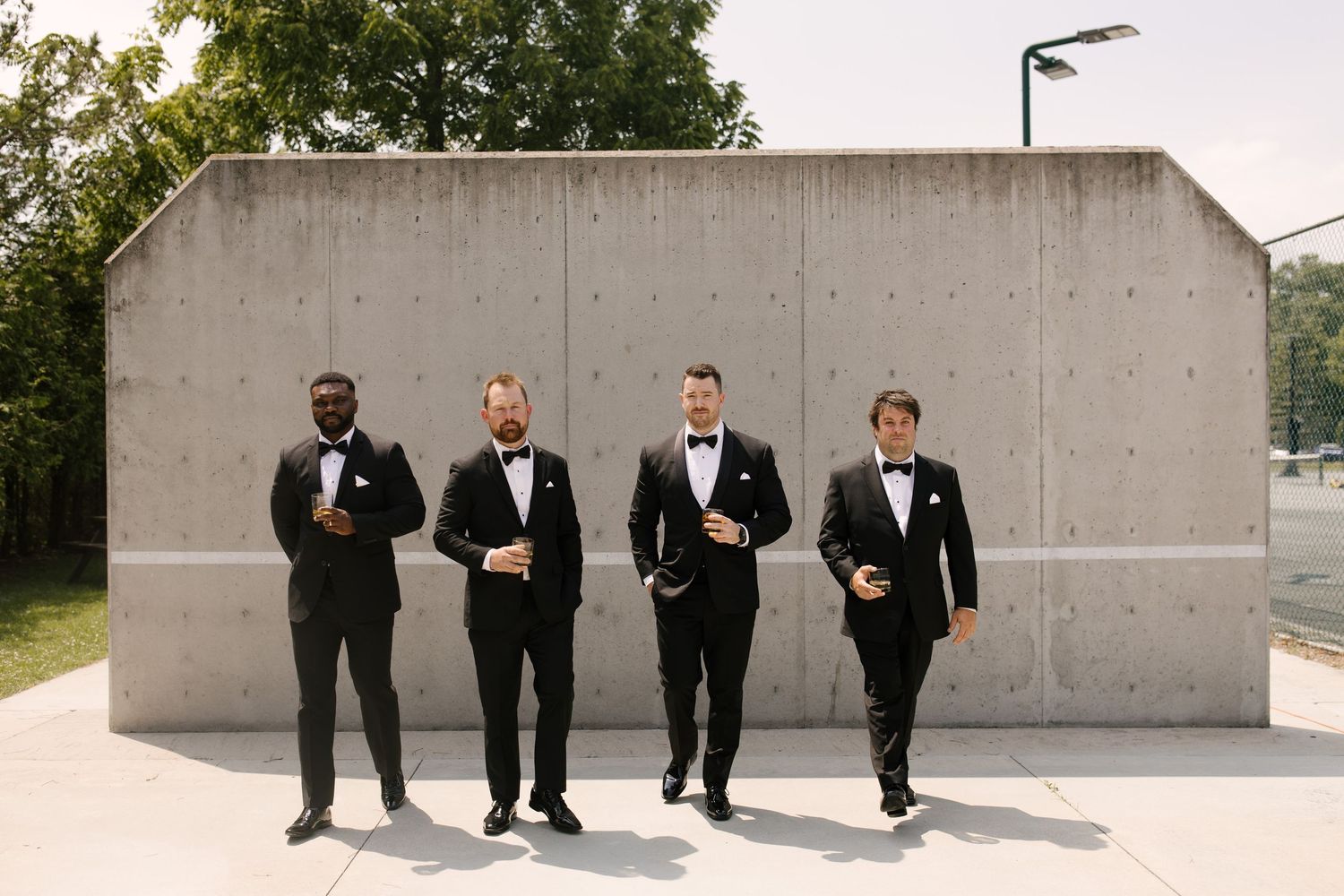 Four groomsmen in black tuxedos standing confidently in front of modern concrete wall outdoors.