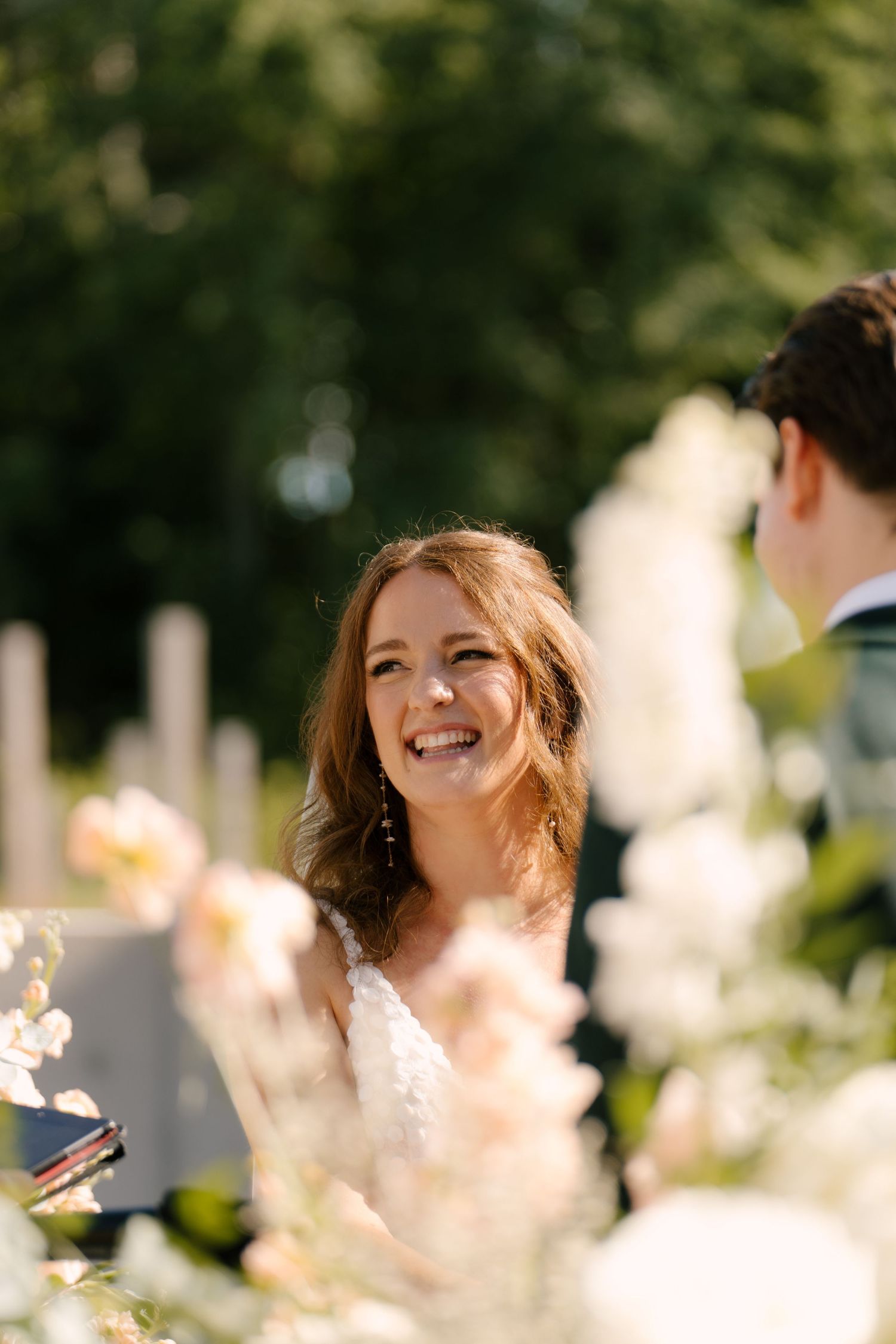 Radiant bride laughing joyfully during outdoor wedding ceremony surrounded by white floral arrangements at Wakefield Estates venue.