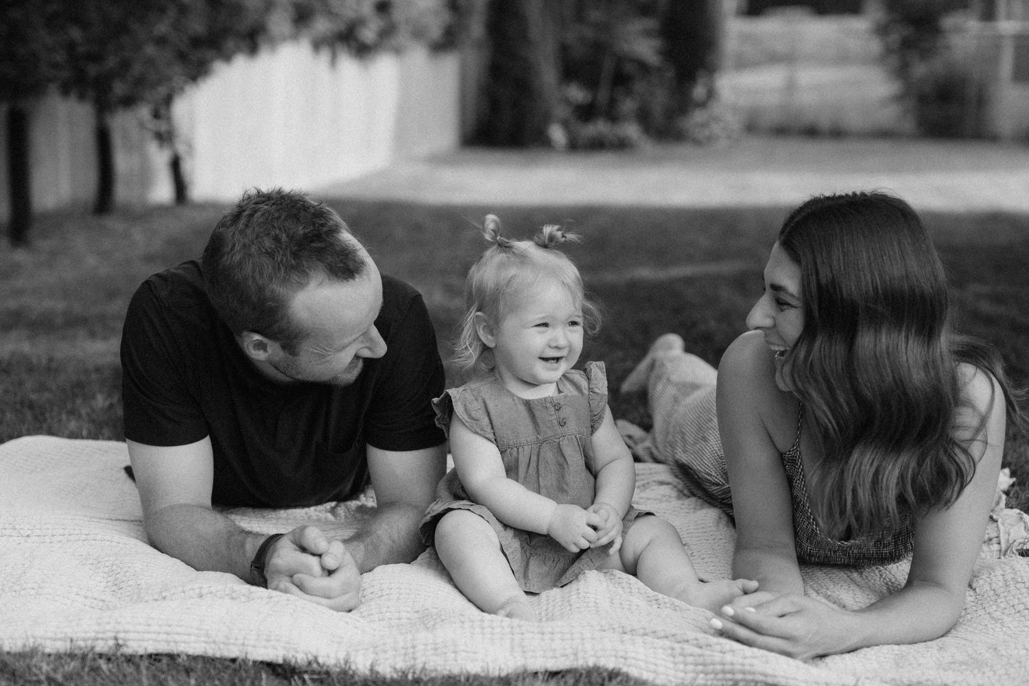 Black and white photo of parents lying on a blanket outdoors smiling at their laughing toddler daughter.