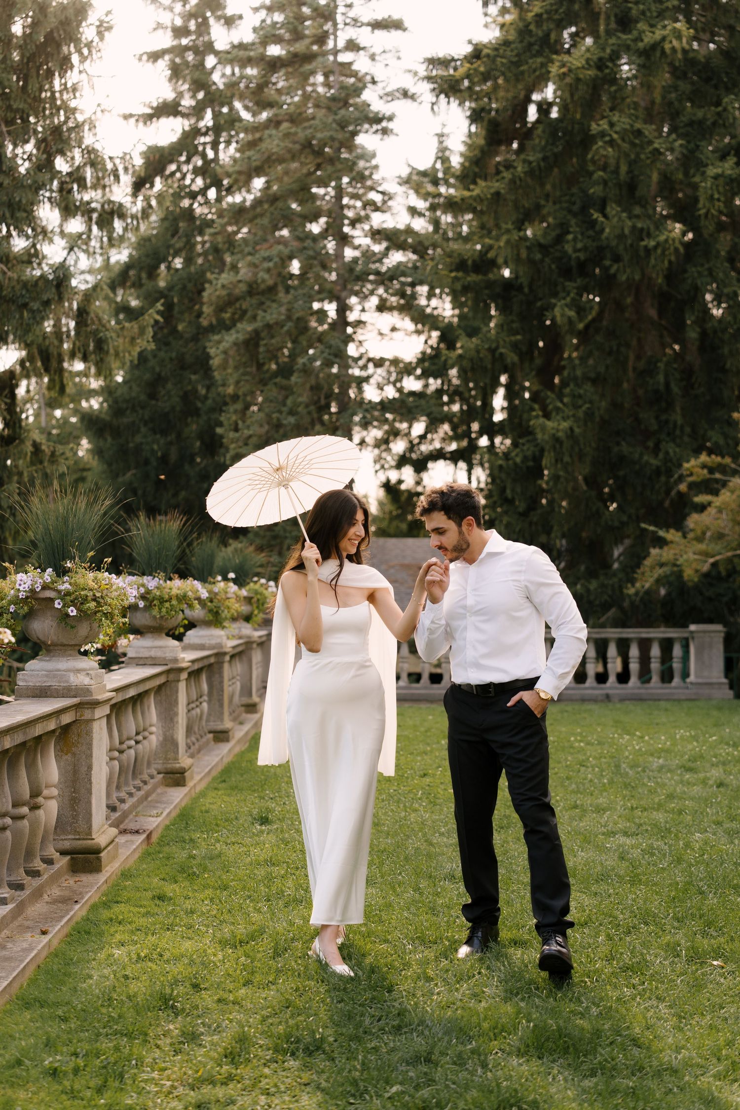 Elegant couple in white dress with parasol and white shirt strolling through a lush garden with stone balustrade.