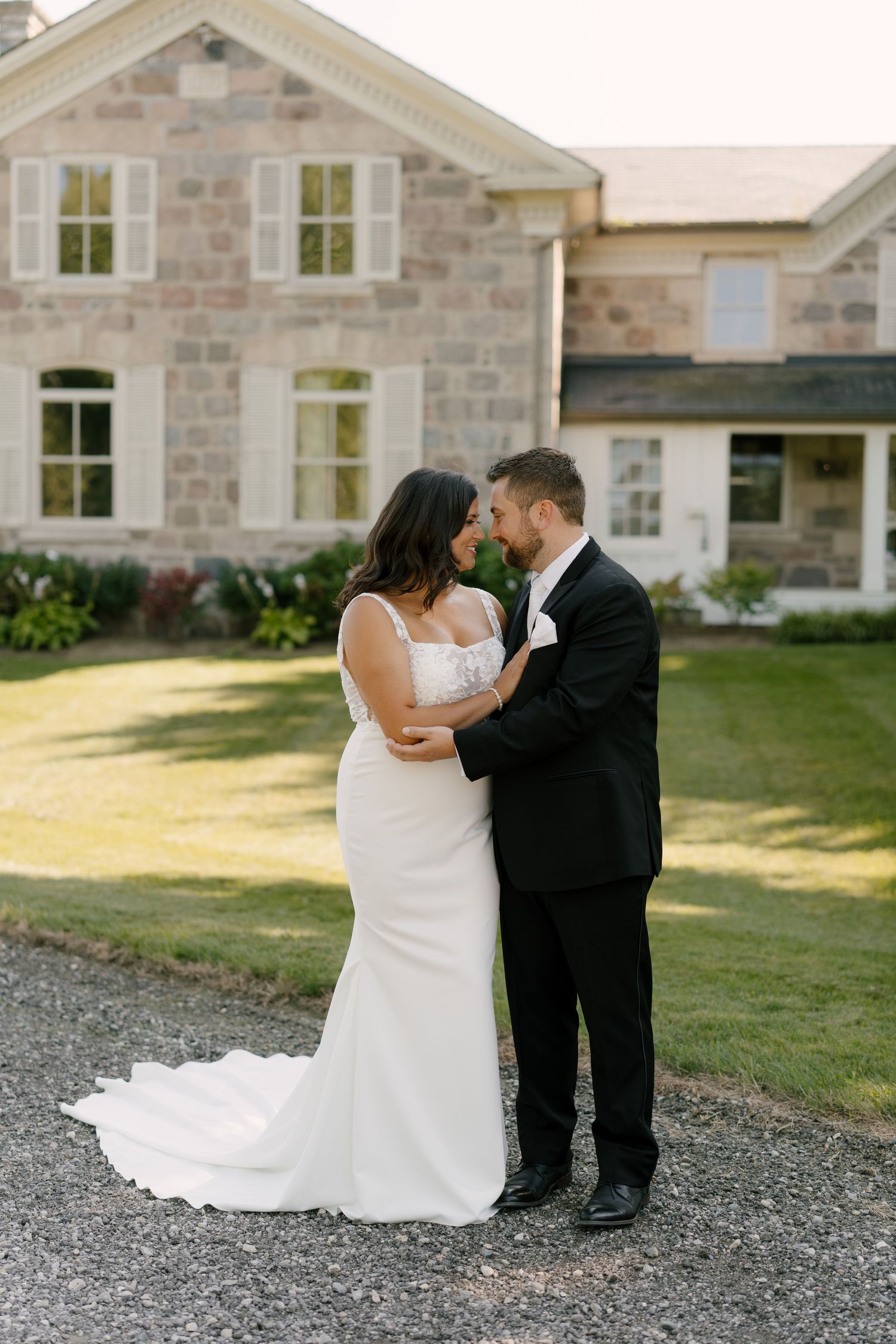 Bride in fitted white gown and groom in black suit embrace on gravel path before stone manor house at Wakefield Estates.