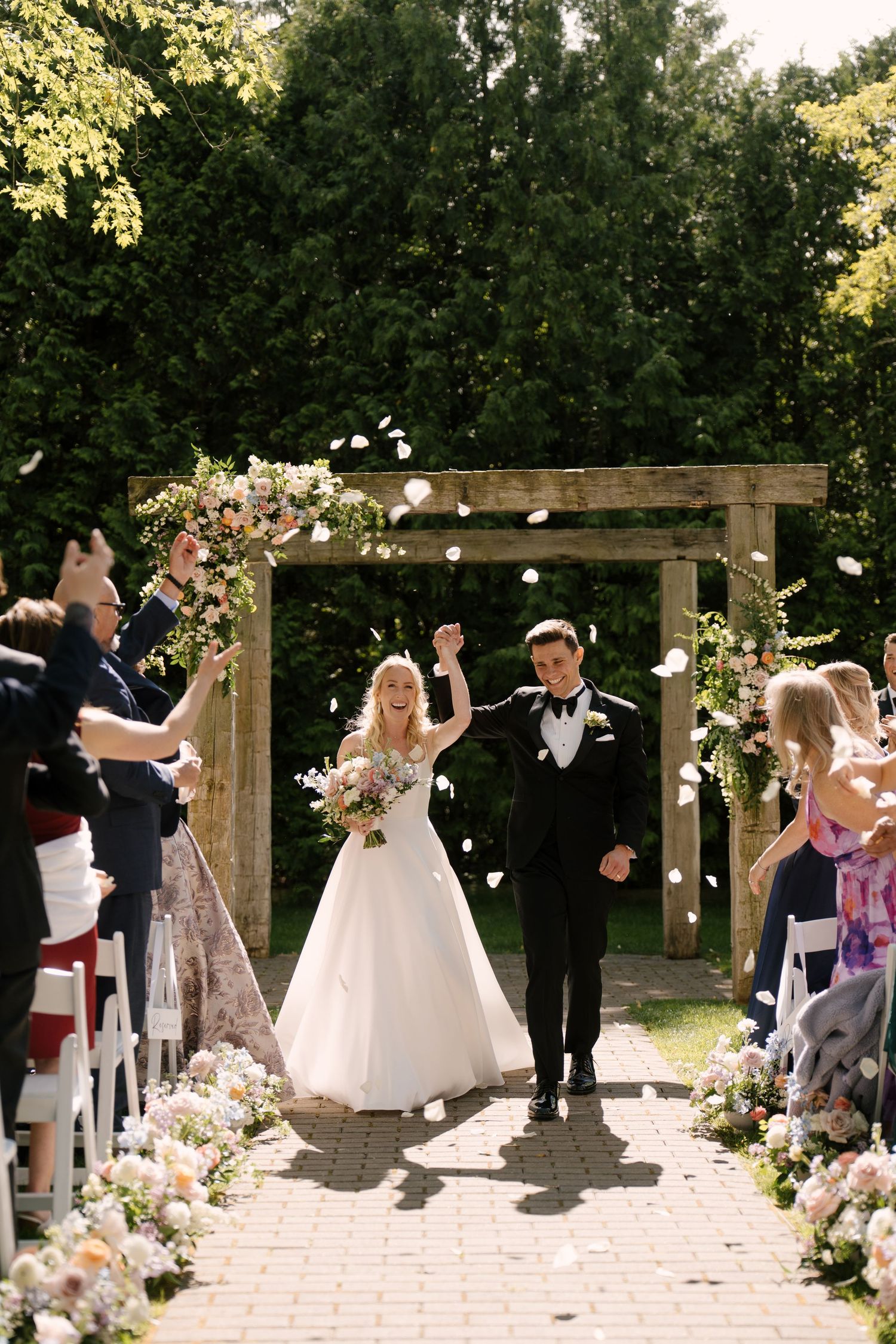 Bride and groom celebrate recessional at outdoor wedding ceremony under wooden arch with floral arrangements.