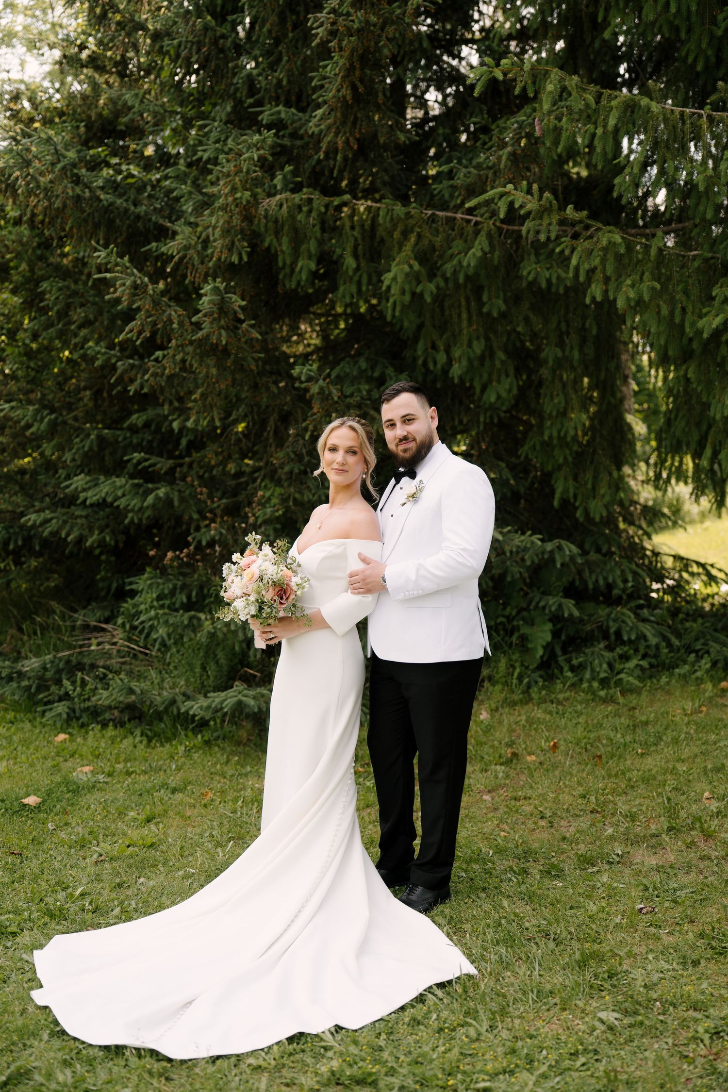 Bride and groom posing together in front of tall evergreen trees, bride holding a soft pastel bouquet.