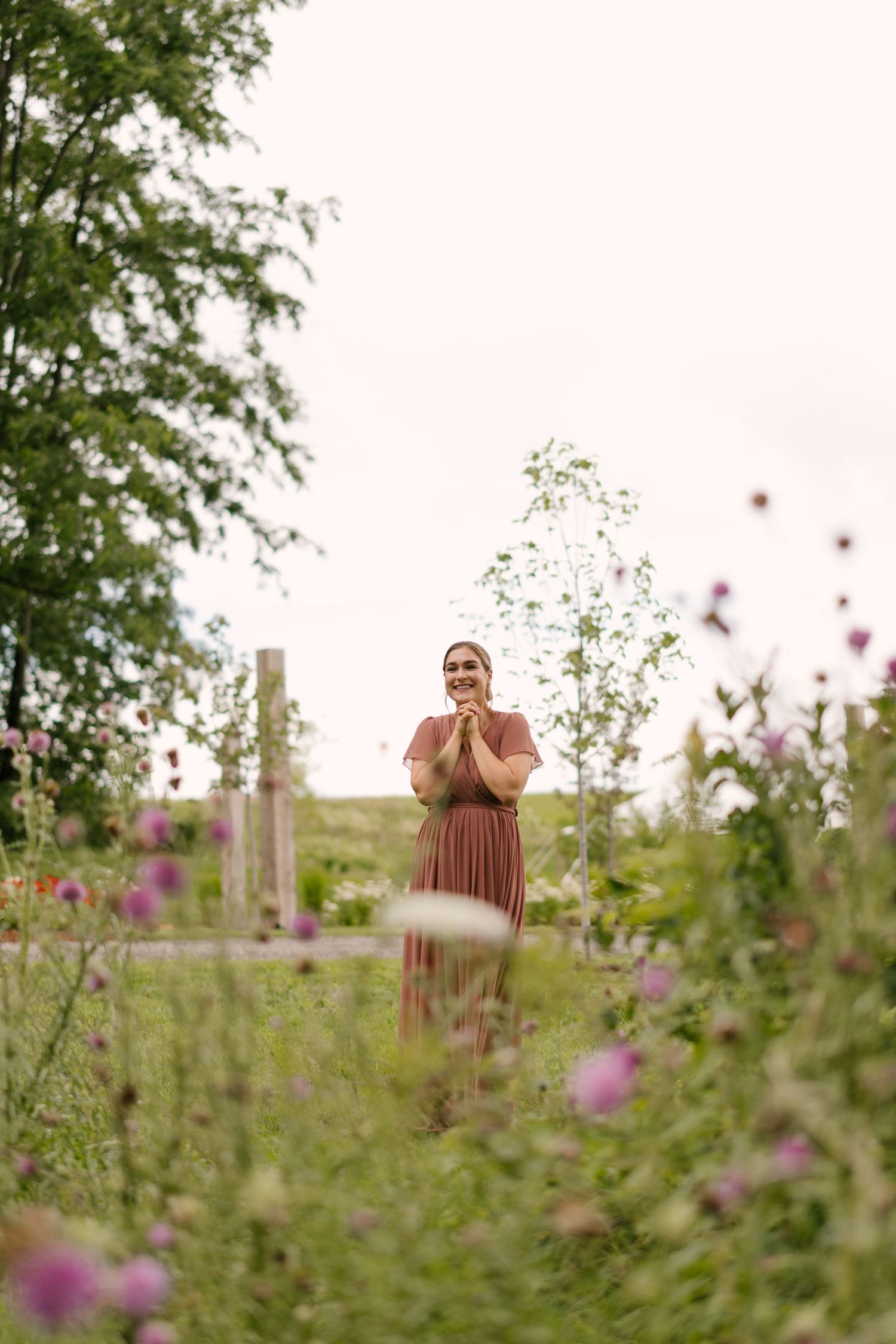 Joyful bridesmaid in mauve dress clasping hands together in wildflower garden setting.
