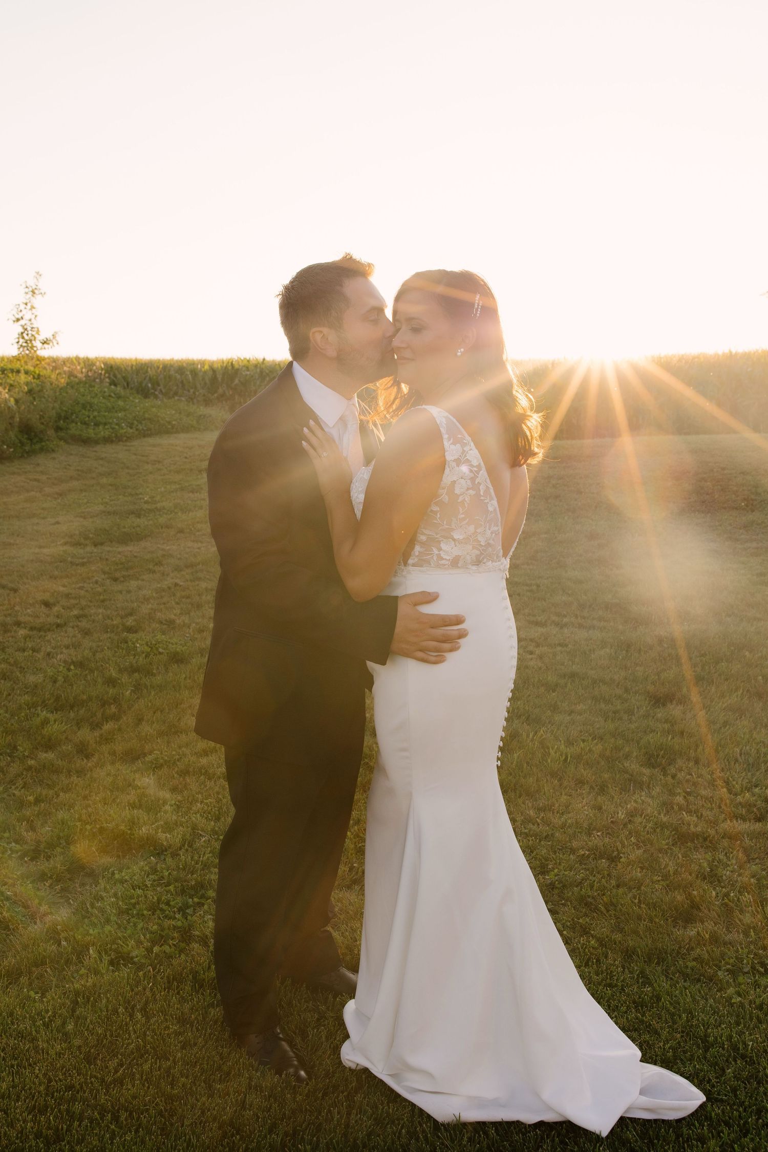 Bride and groom sharing a romantic kiss at golden hour sunset on a lush green lawn.