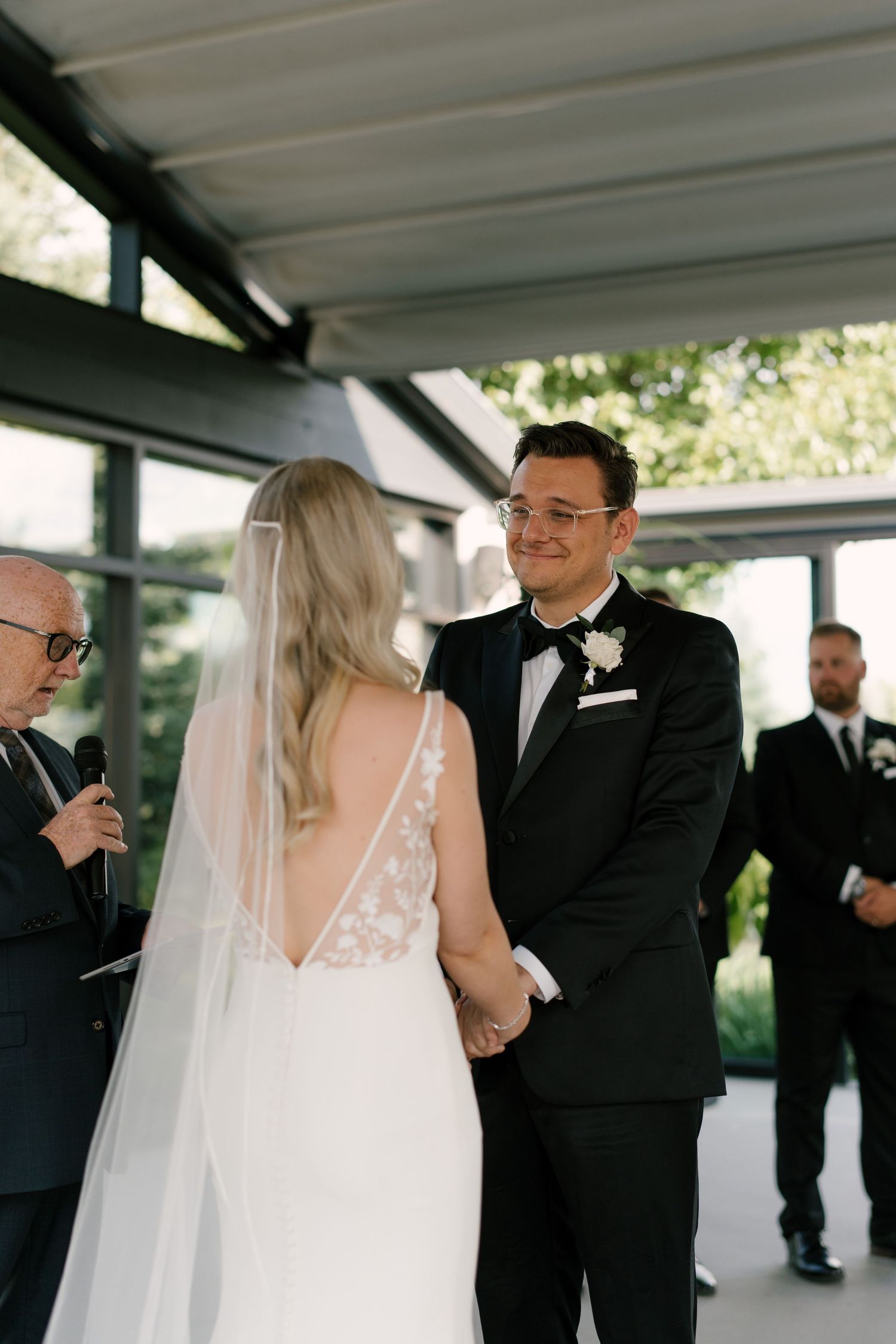 Groom in a black tuxedo smiling at his bride during an outdoor wedding ceremony while holding hands.