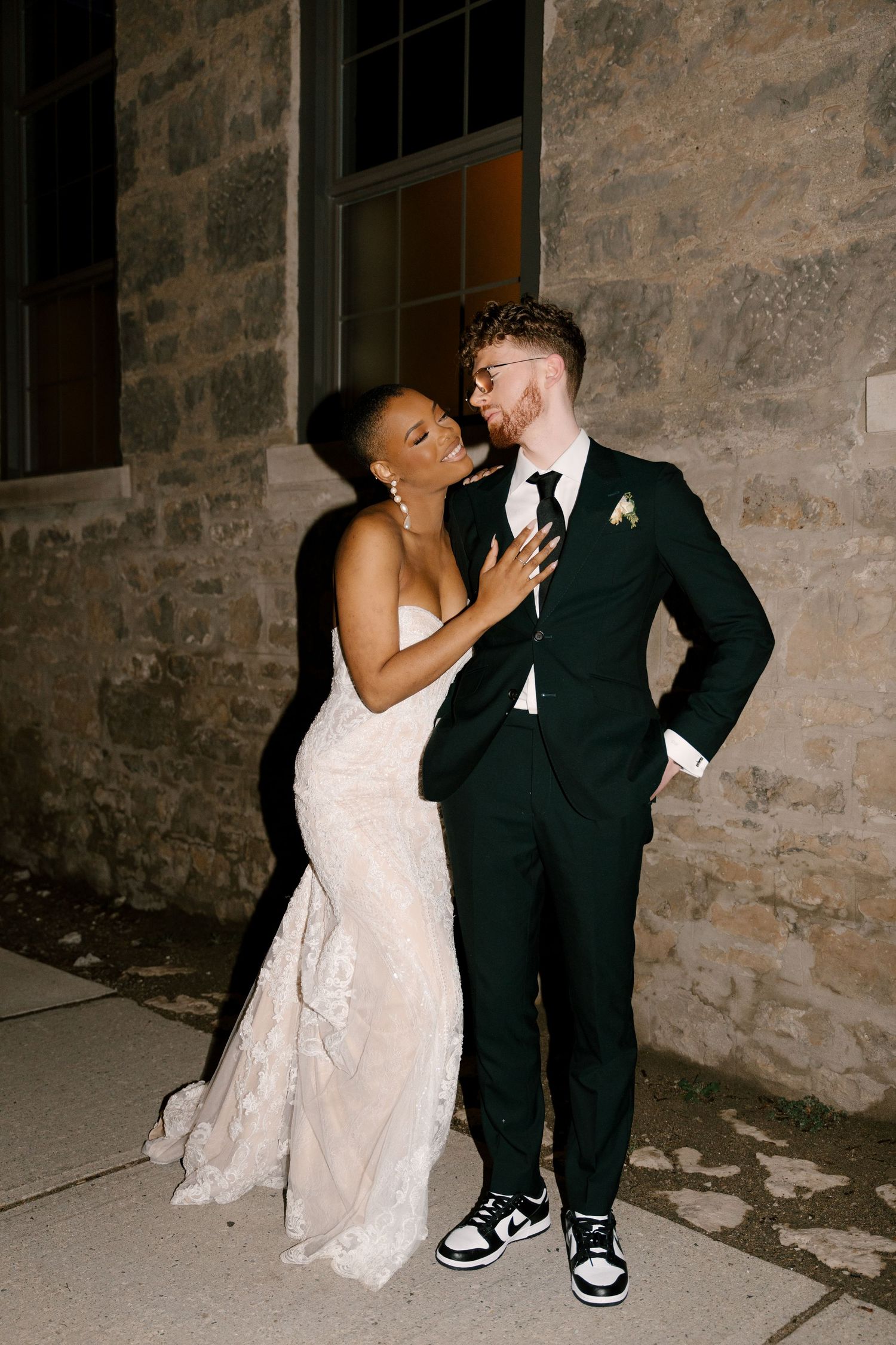 Romantic newlywed couple sharing an intimate moment outside against a stone wall at night.