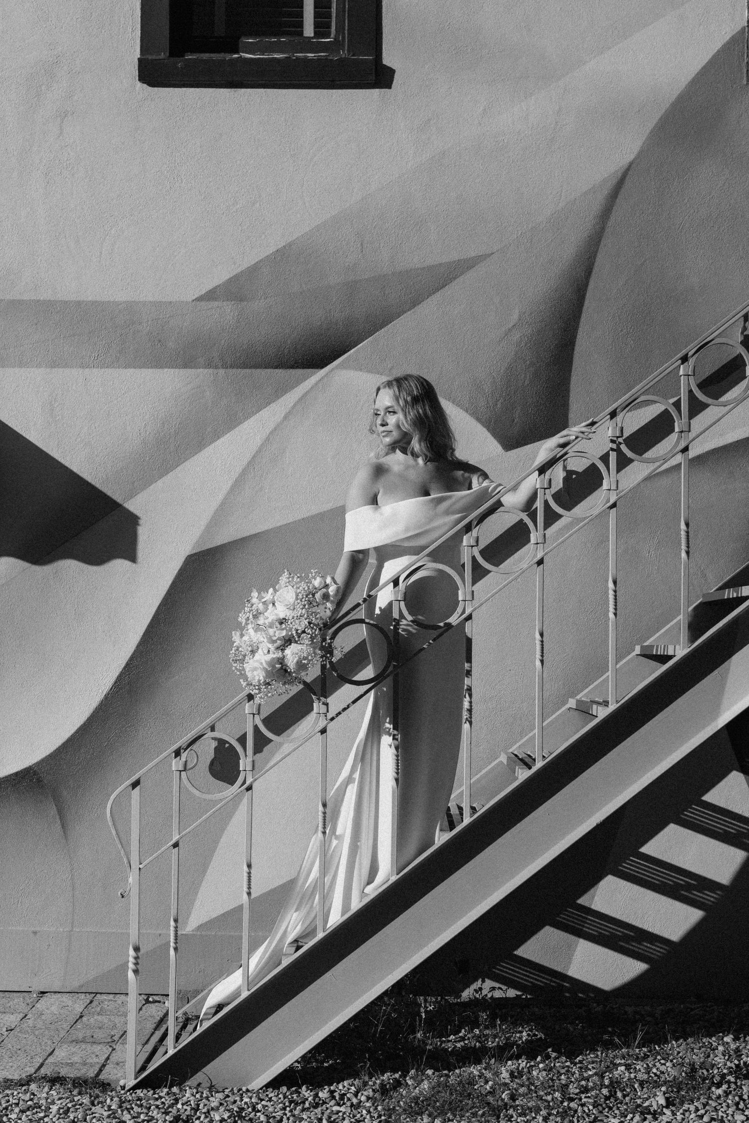 Black and white photo of a radiant bride in an off-shoulder gown descending an outdoor staircase with bouquet.