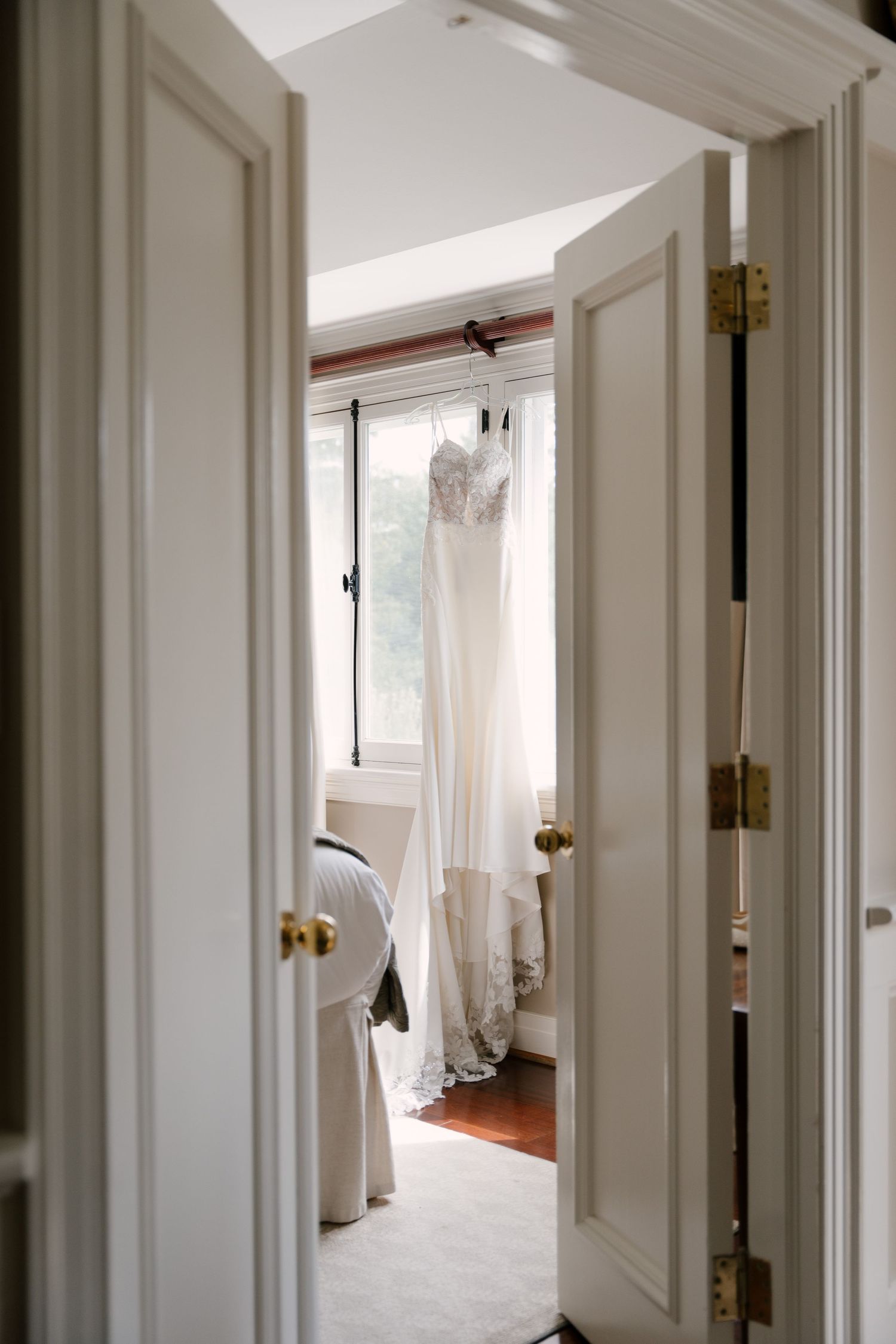 A white wedding dress hanging in a sunlit bedroom doorway with elegant double doors and gold hinges.