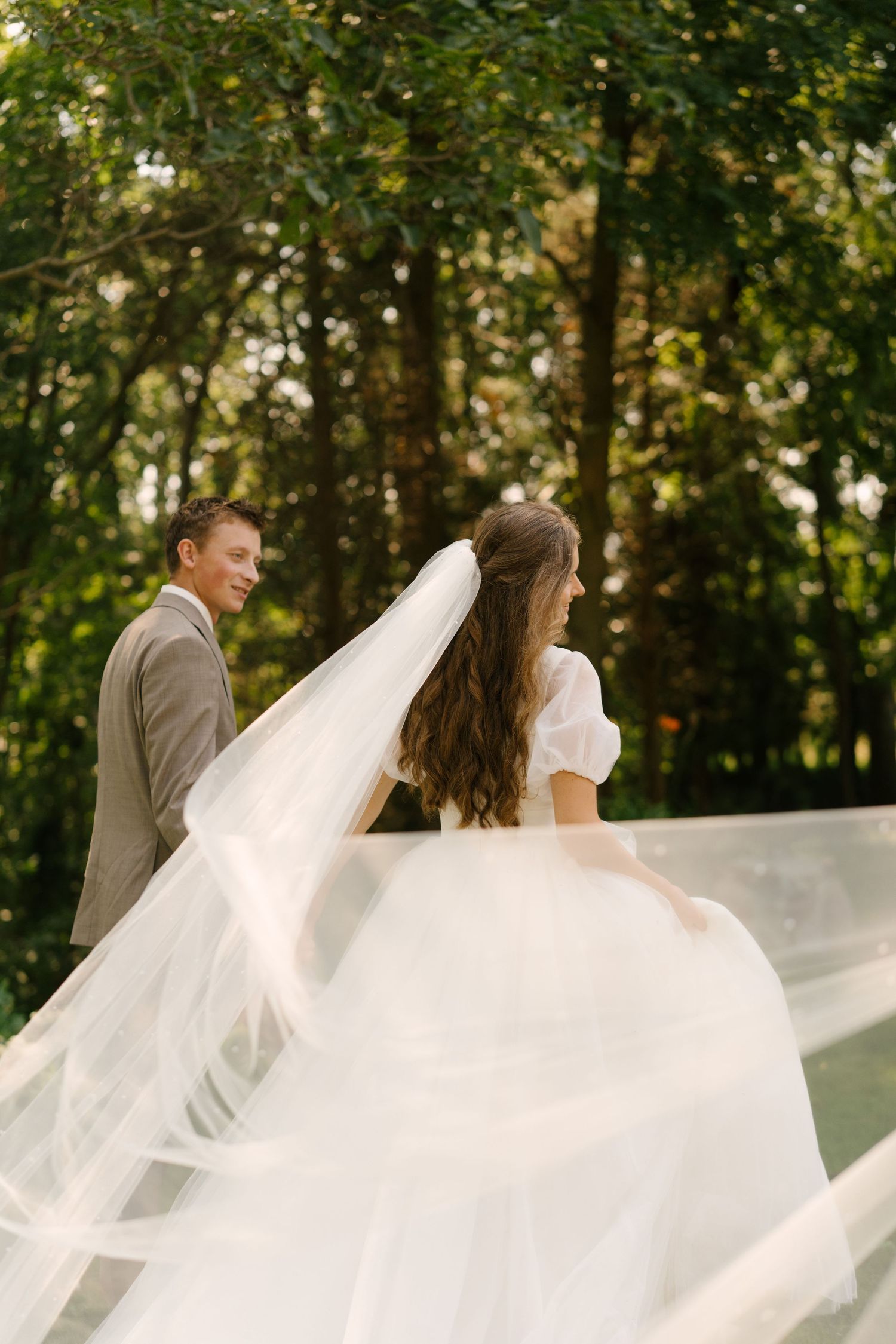 Smiling groom watches bride as her white veil and gown flow dramatically in a sunlit forest setting.