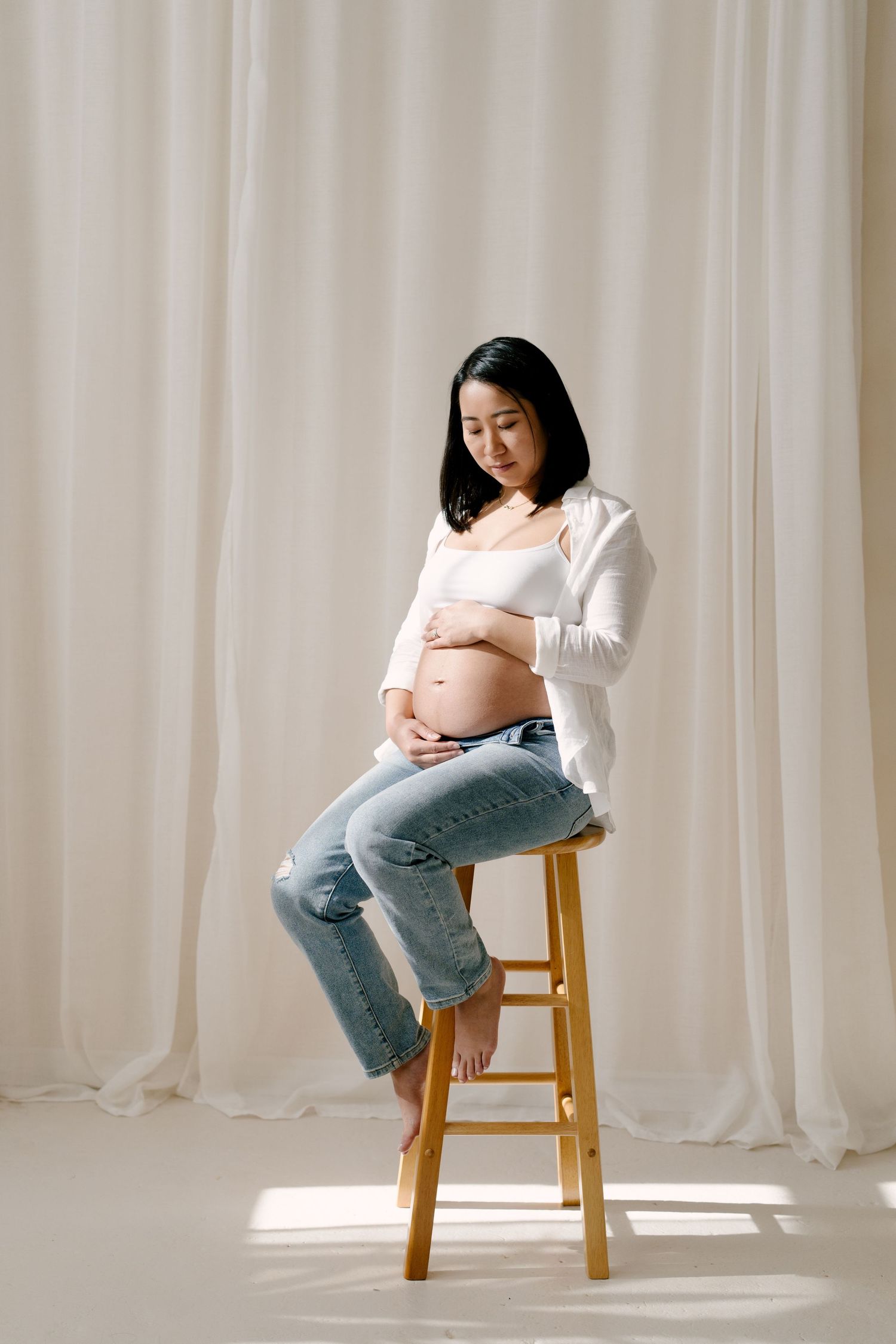 Pregnant woman in white shirt and jeans sits on wooden stool, cradling bump, before white curtains.
