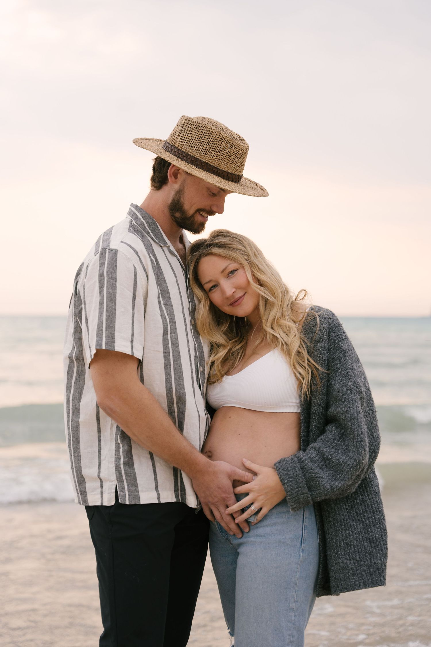 Couple at beach sunset, man in straw hat embracing pregnant woman holding her baby bump.