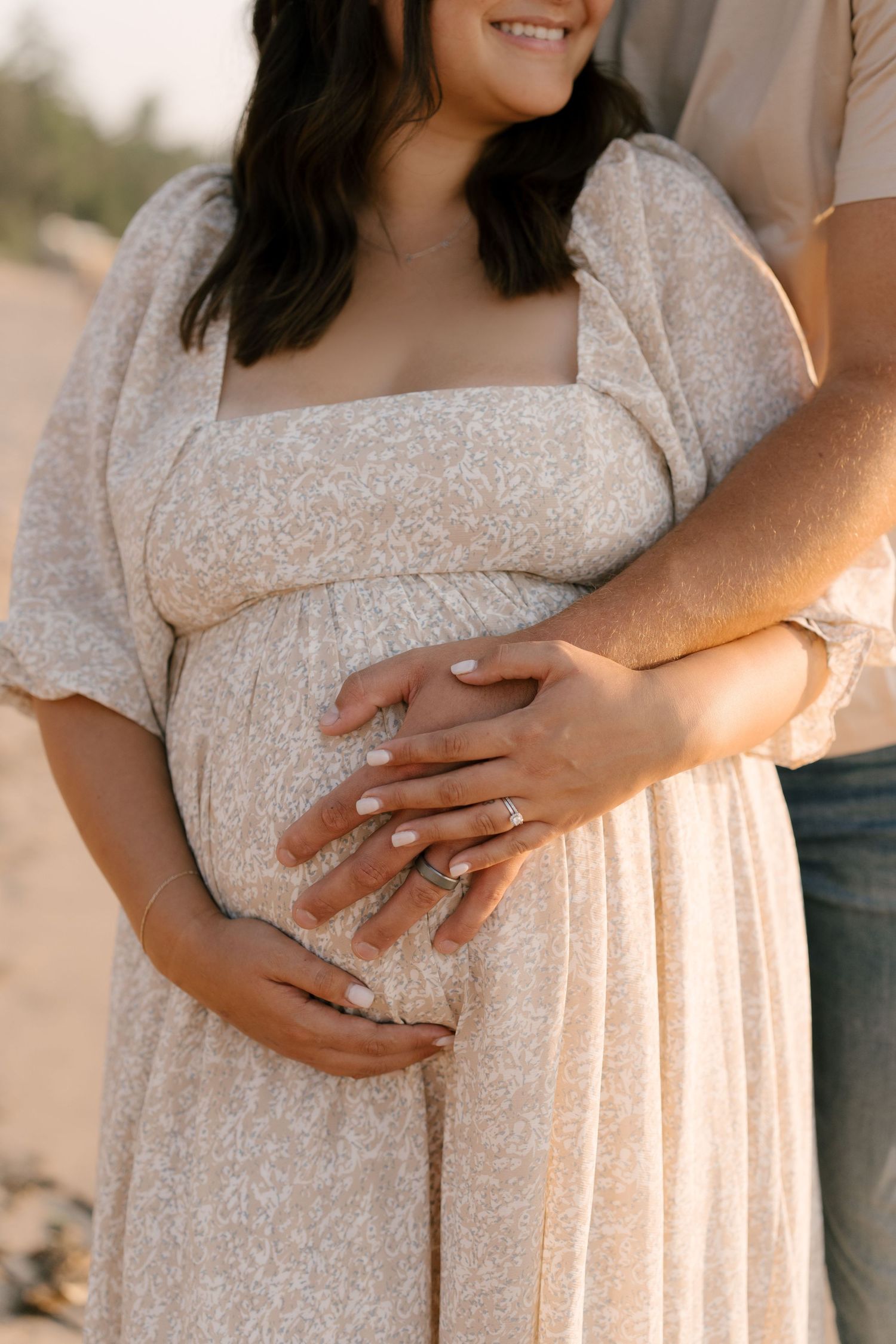Close-up of pregnant womans baby bump with couples hands resting on white floral maternity dress.