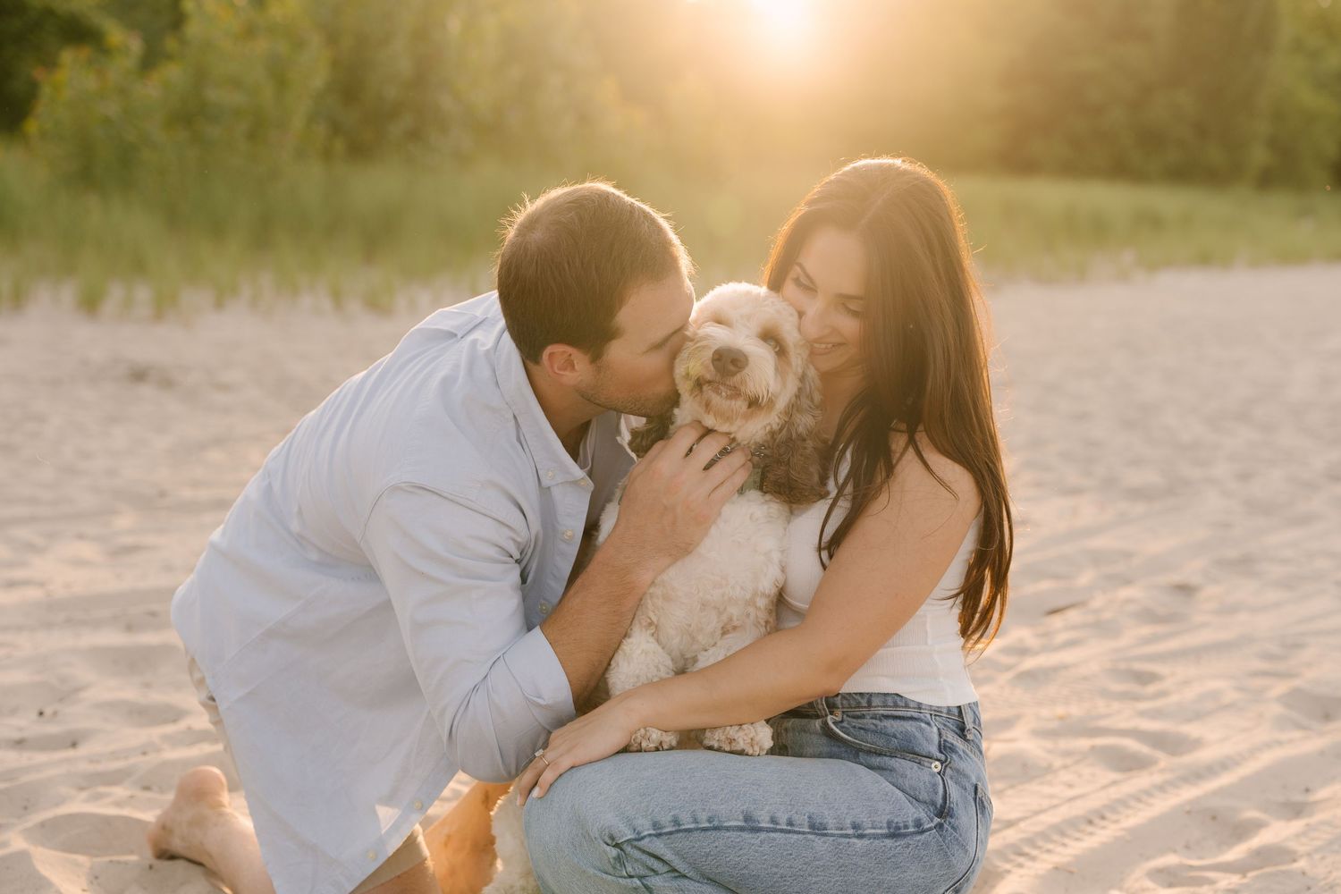 Couple kissing their fluffy white dog on sandy beach at warm golden sunset light.