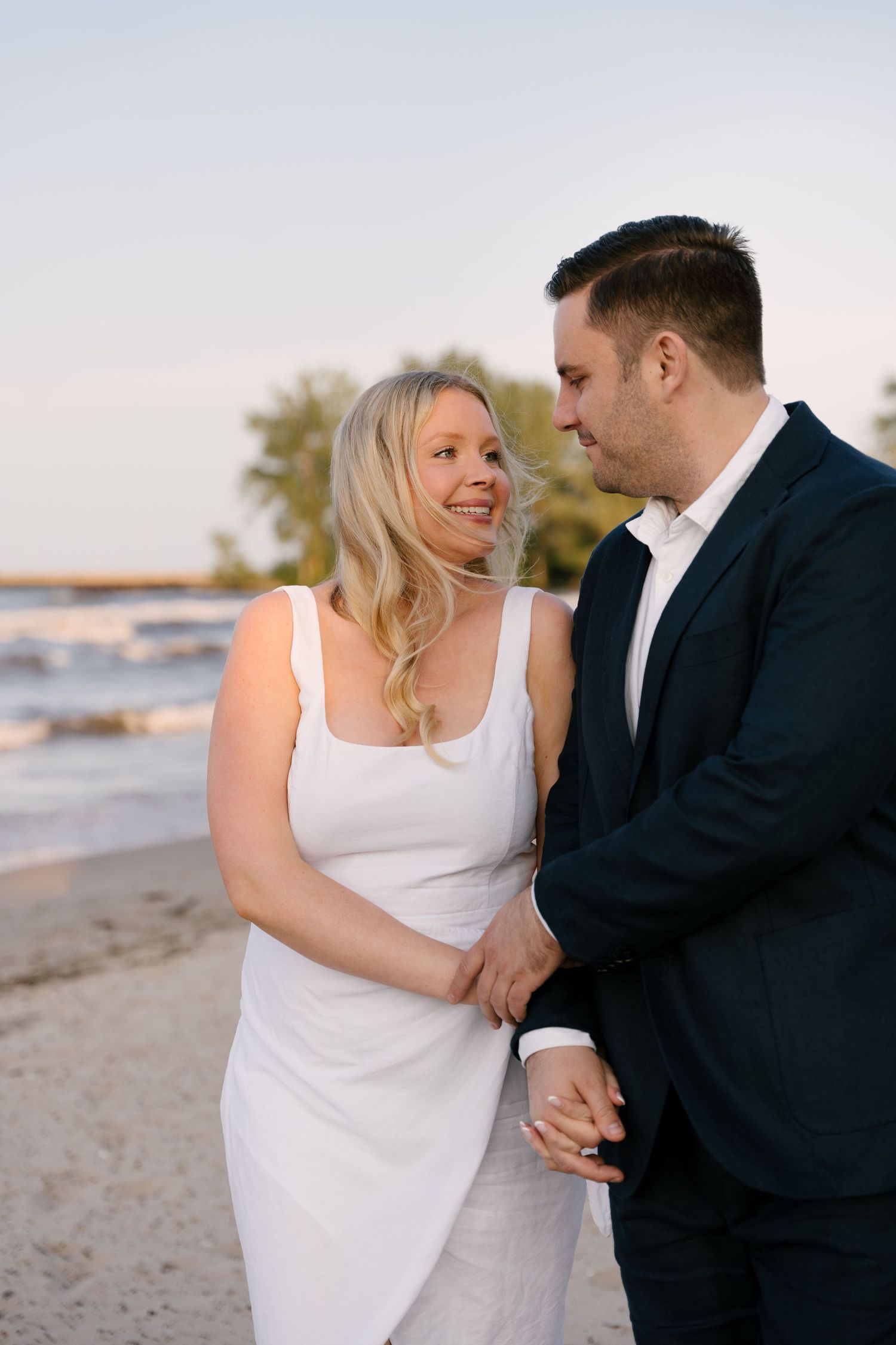 Engaged couple holding hands and smiling at each other on a sandy beach at sunset.