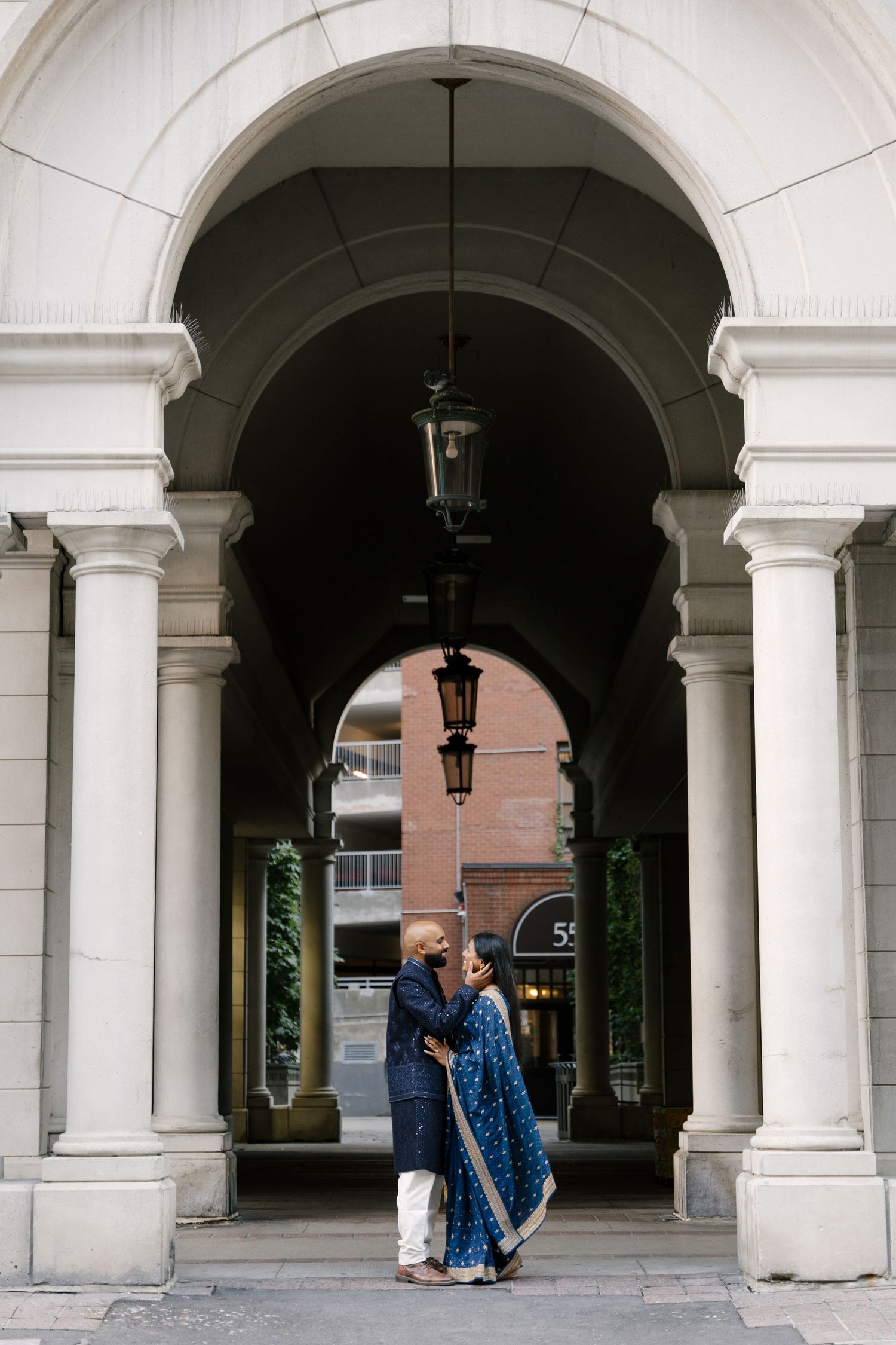 Couple in traditional Indian attire embracing under elegant white arched colonnade with hanging lanterns.