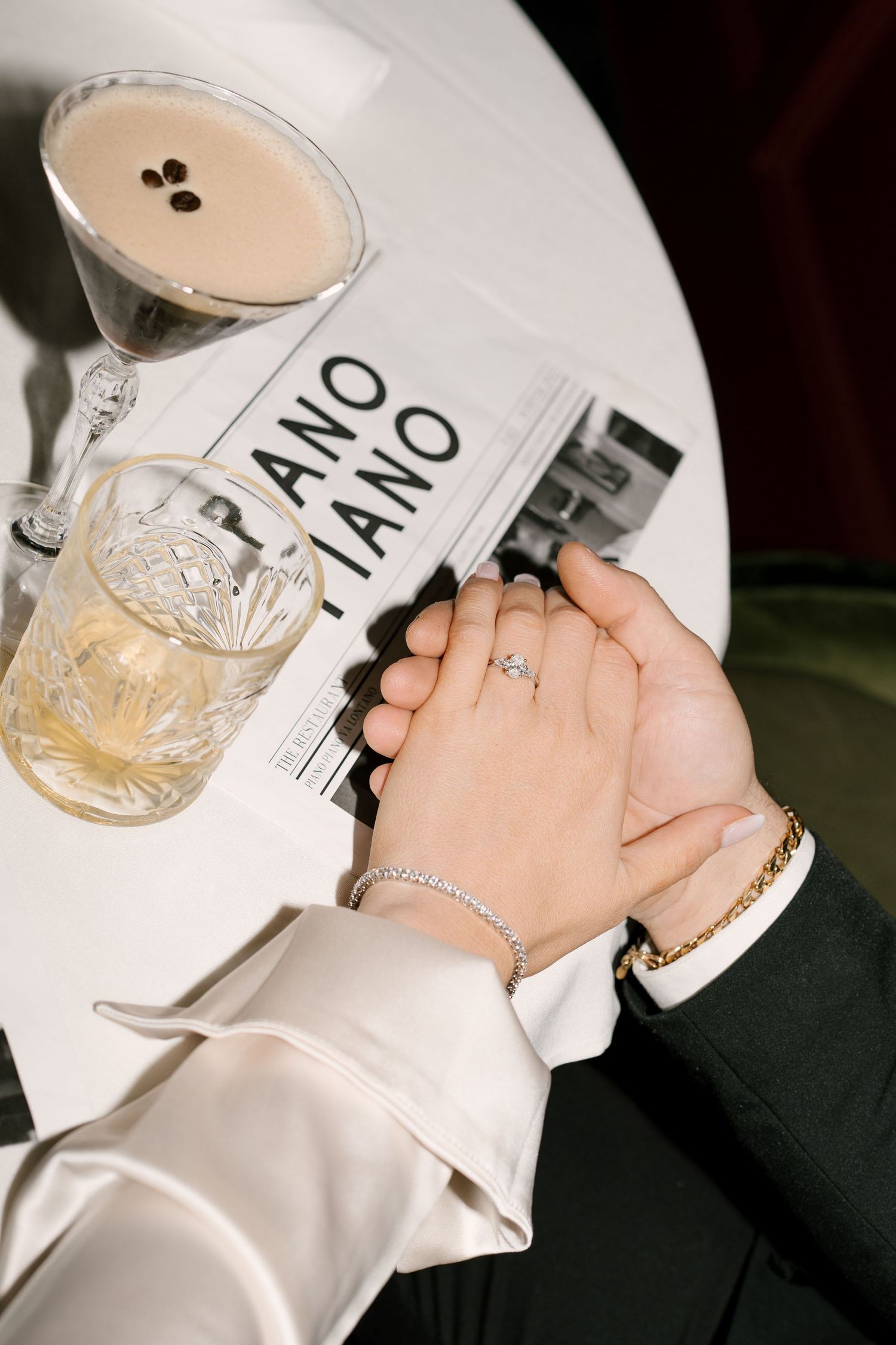 Close-up of engaged couple holding hands showing diamond ring at Piano Piano restaurant table with cocktails.