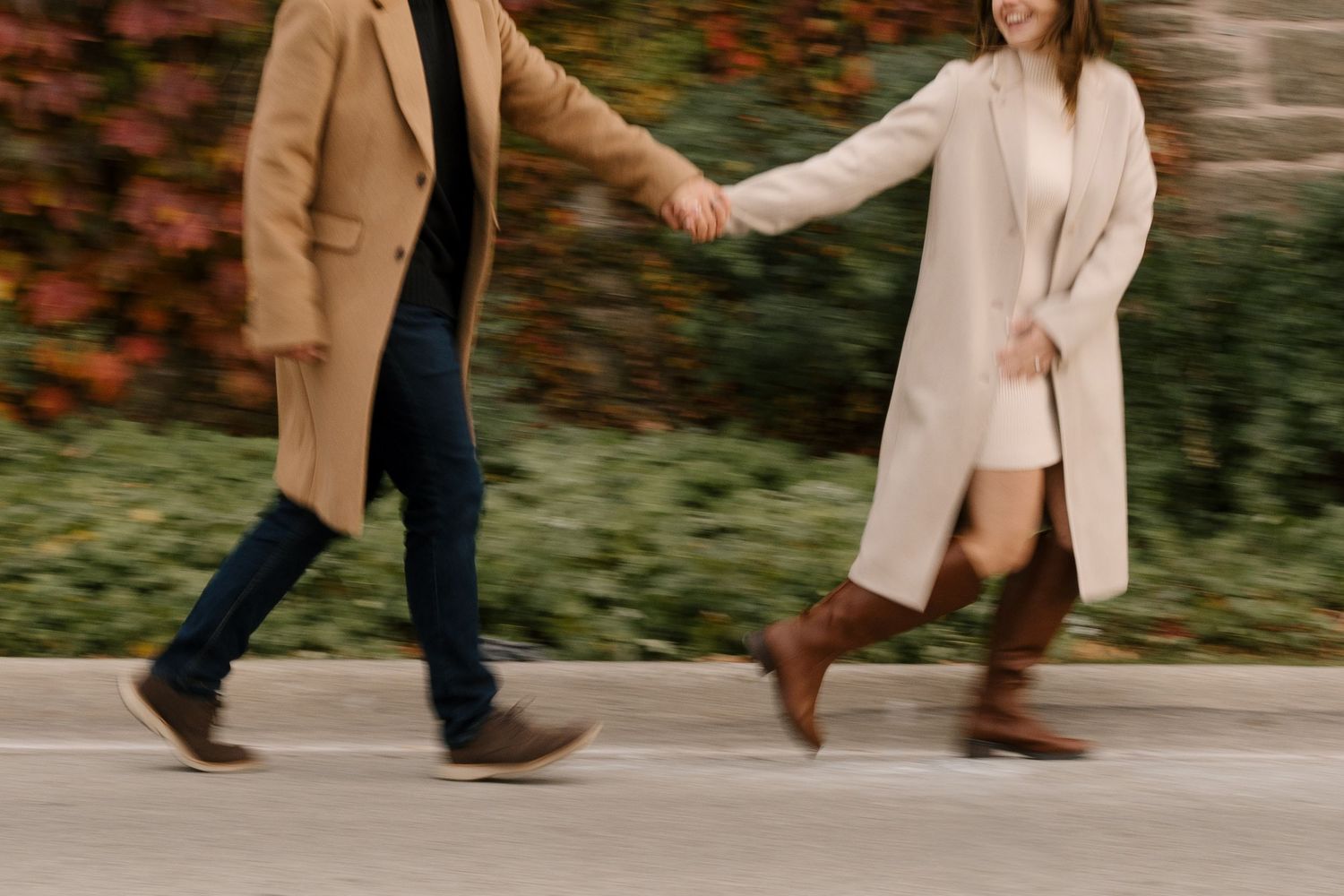Motion-blurred couple holding hands walking briskly past fall foliage in coordinating camel coats.